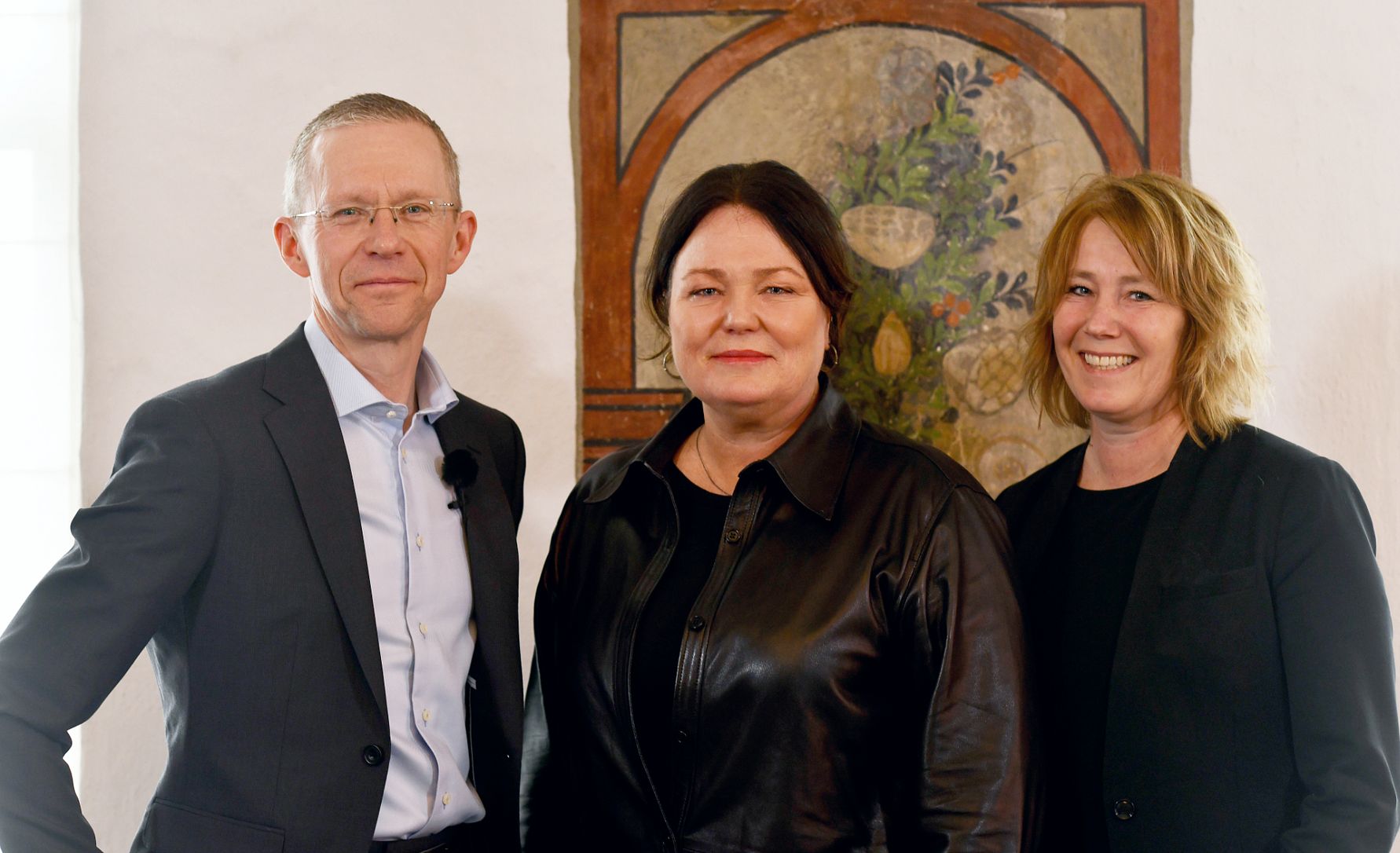 Tre personer står framför en målning med växtmotiv, iförda formella kläder