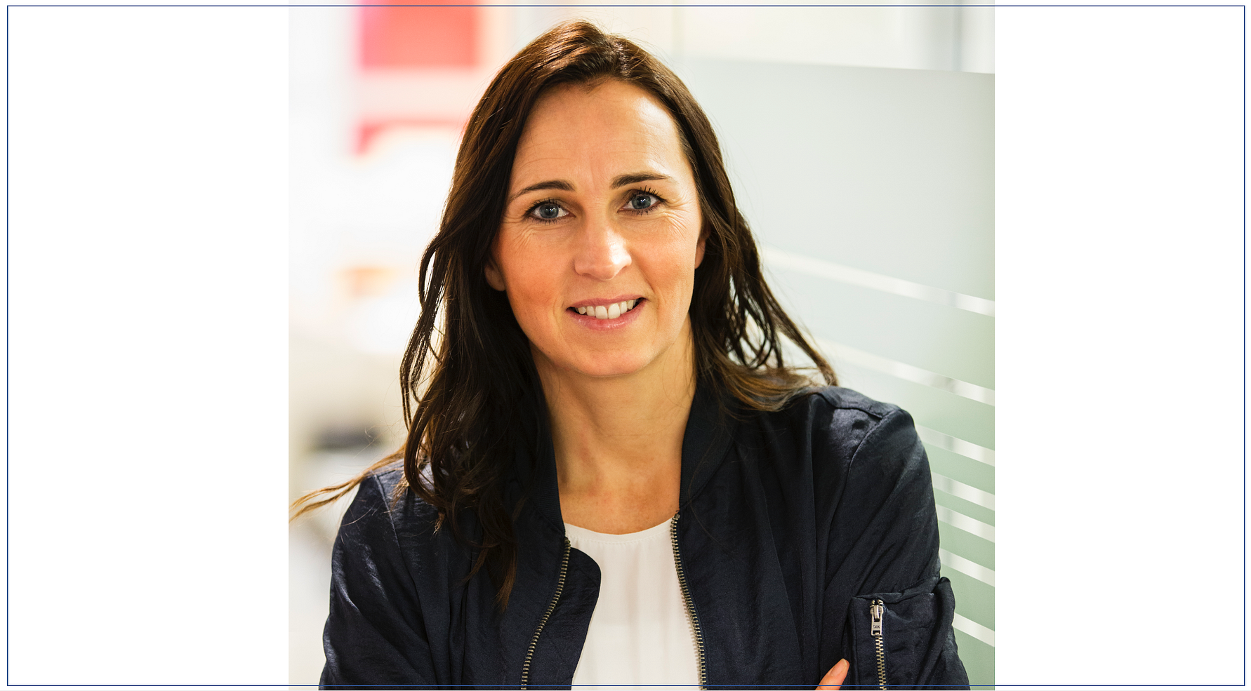 En kvinna med långt mörkt hår iförd en mörkblå jacka och en vit topp står med korsade armar framför en glasvägg i en ljus inomhusmiljö