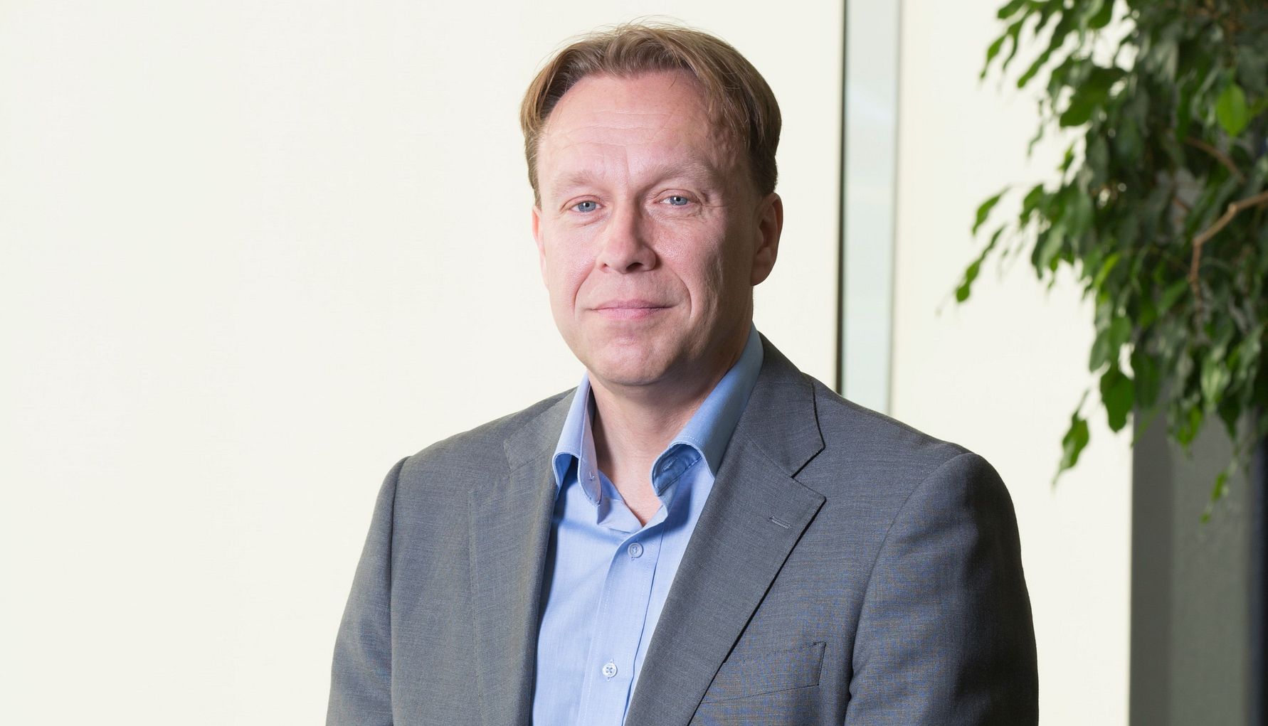 Man i grå kavaj och ljusblå skjorta står framför en ljus bakgrund med en grön växt i högerkant