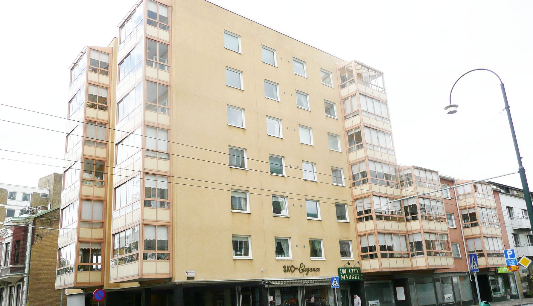 Flerbostadshus med beige fasad och stora fönster, beläget vid en gata med affärer på bottenvåningen