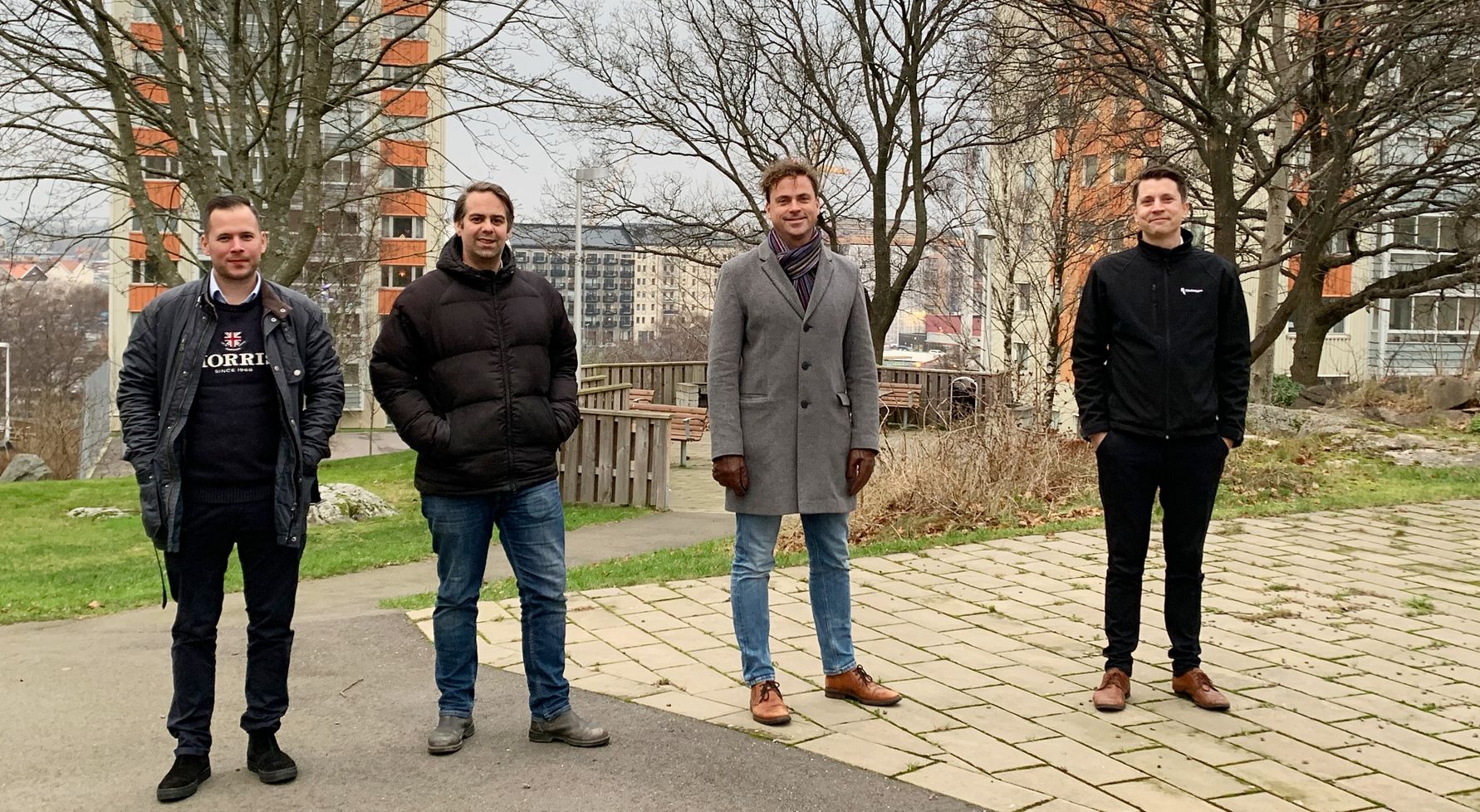 Fyra personer står utomhus i en urban miljö med gräsmatta, träd, parkbänk och flerfamiljshus i bakgrunden
