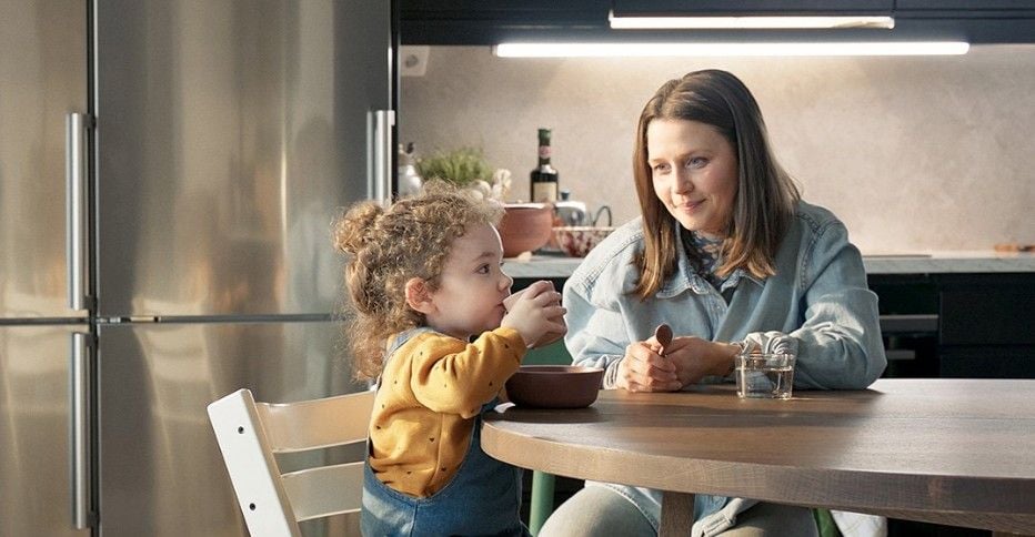 En vuxen och ett barn sitter vid ett runt matbord i ett modernt kök med rostfri kyl och mörka skåp i bakgrunden