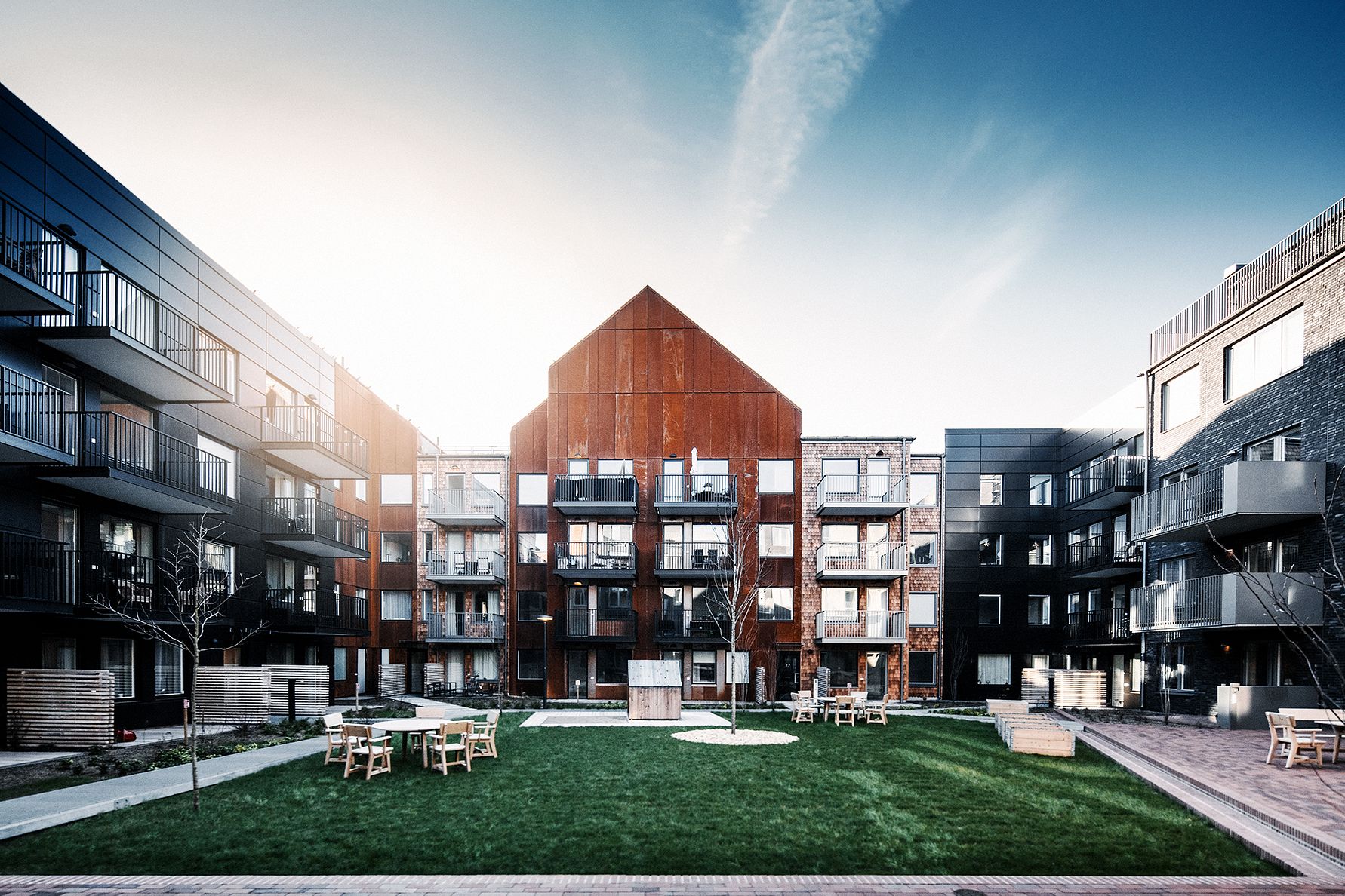 Modern bostadsområde med en central byggnad i rödbrunt trä omgiven av svarta flerfamiljshus, grön gräsmatta, och sittmöbler i en gemensam gård under en klar himmel
