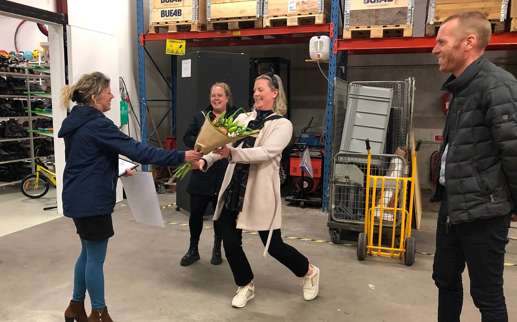 Person tar emot en blombukett på ett lagergolv omgivet av hyllor, kartonger och en gul vagn