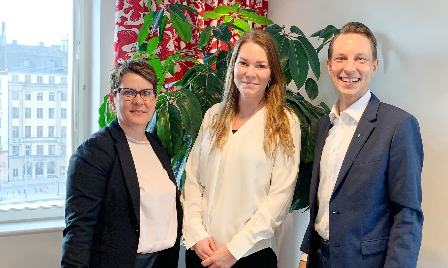 Tre personer står inomhus framför en stor grön växt och ett fönster med röd-vit gardin. Bakgrunden visar en stadsbild med byggnader