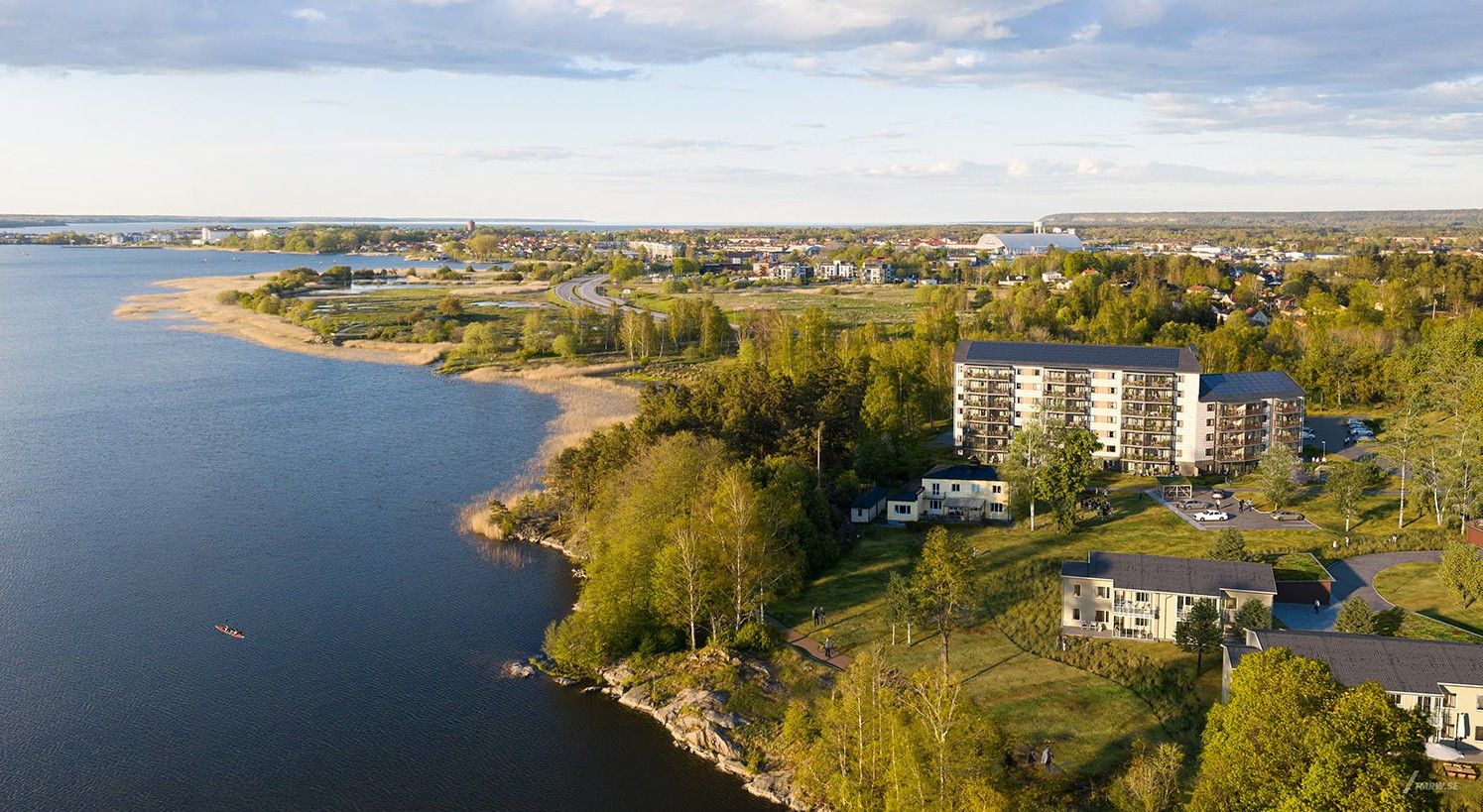 Flygfoto av ett bostadsområde nära en sjö med omgivande grönska och utsikt över en stad i fjärran
