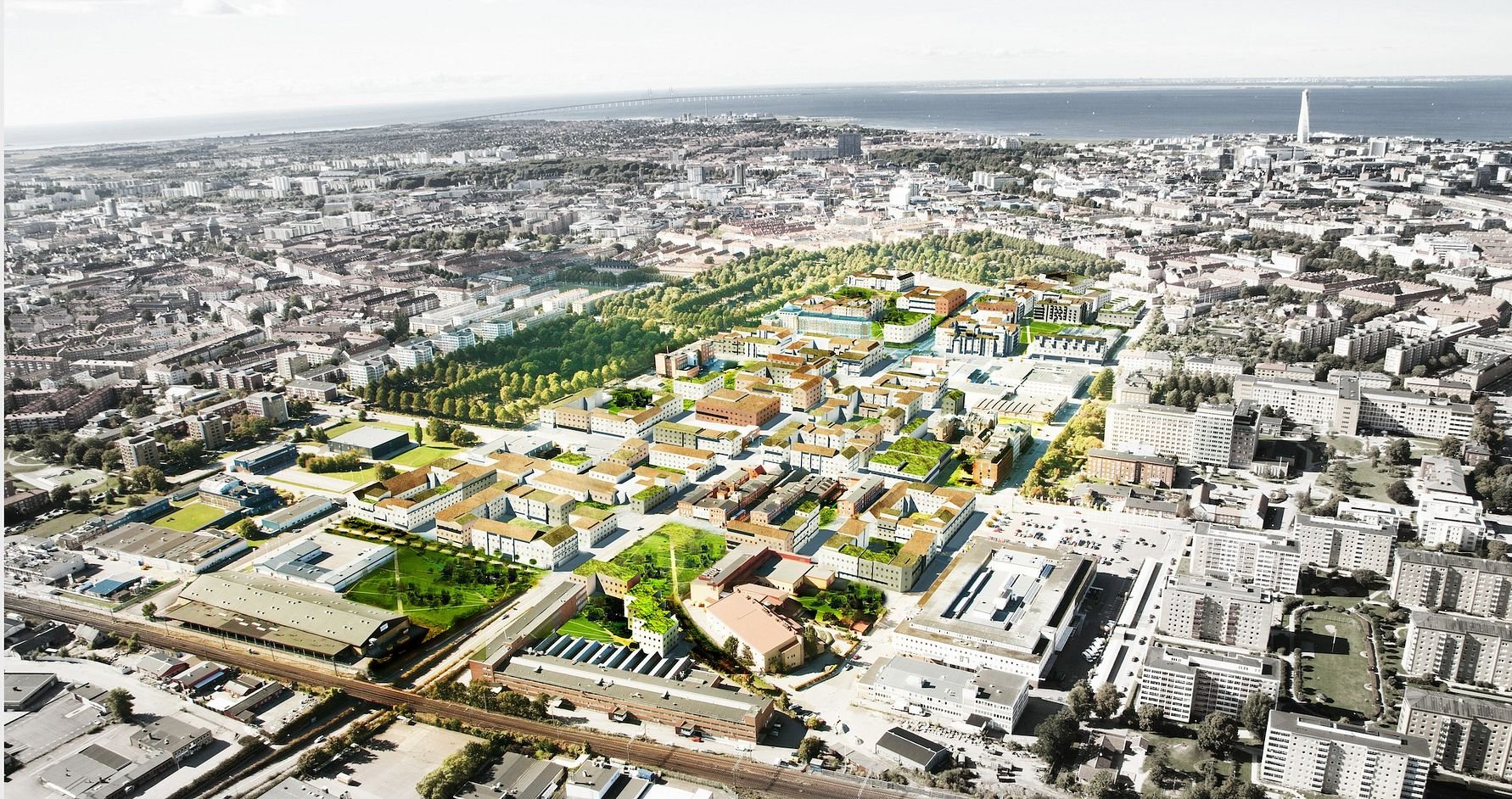 Flygfoto över en stadsdel med en blandning av byggnader, parker och grönområden, samt en kustlinje i bakgrunden