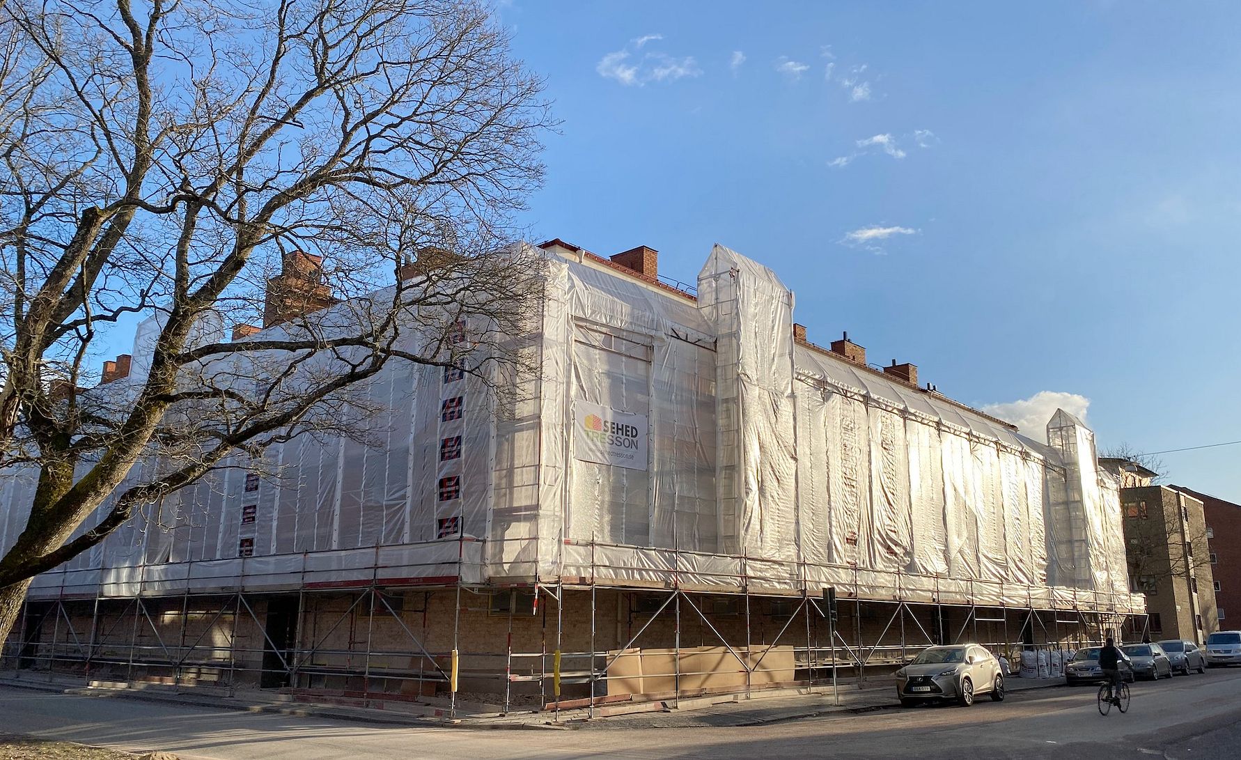 Byggnad täckt av byggnadsställningar och vit skyddsplast, med en klar blå himmel i bakgrunden. En bil och en cyklist syns på gatan framför byggnaden
