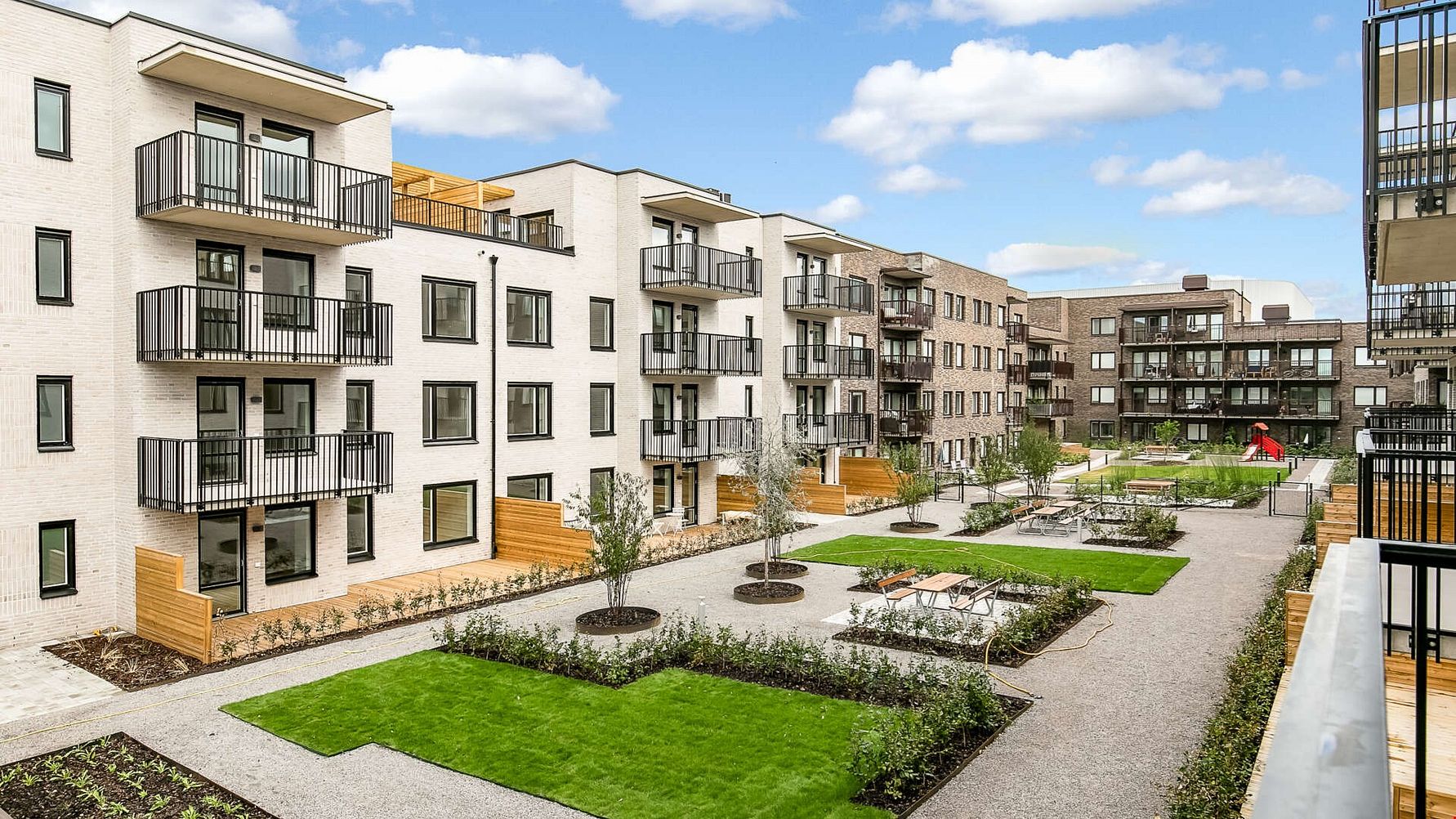 Modern bostadsområde med vita flerfamiljshus, balkonger och välskötta grönområden samt gångvägar under en klar blå himmel