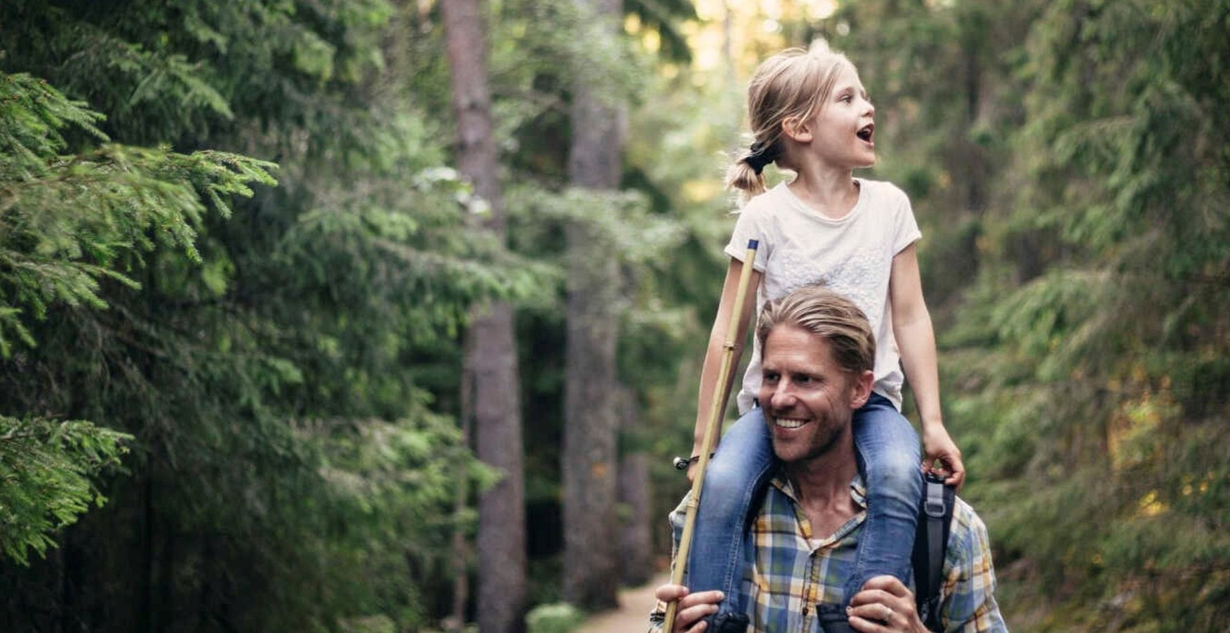 En vuxen person bär ett barn på axlarna medan de promenerar genom en skog med höga träd och grönska