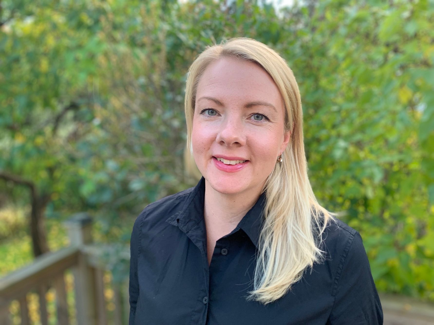 En person med ljust hår och svart skjorta står utomhus med grönskande träd i bakgrunden