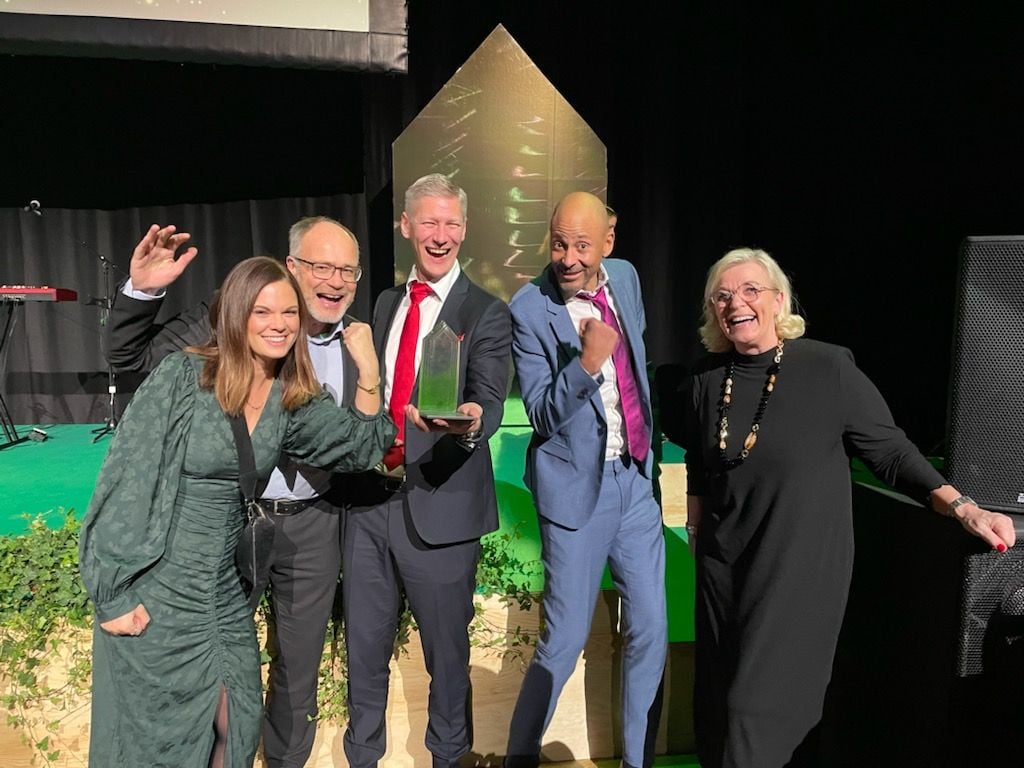 En grupp människor poserar på en scen med en prisstatyett, omgivna av grönska och svart bakgrund