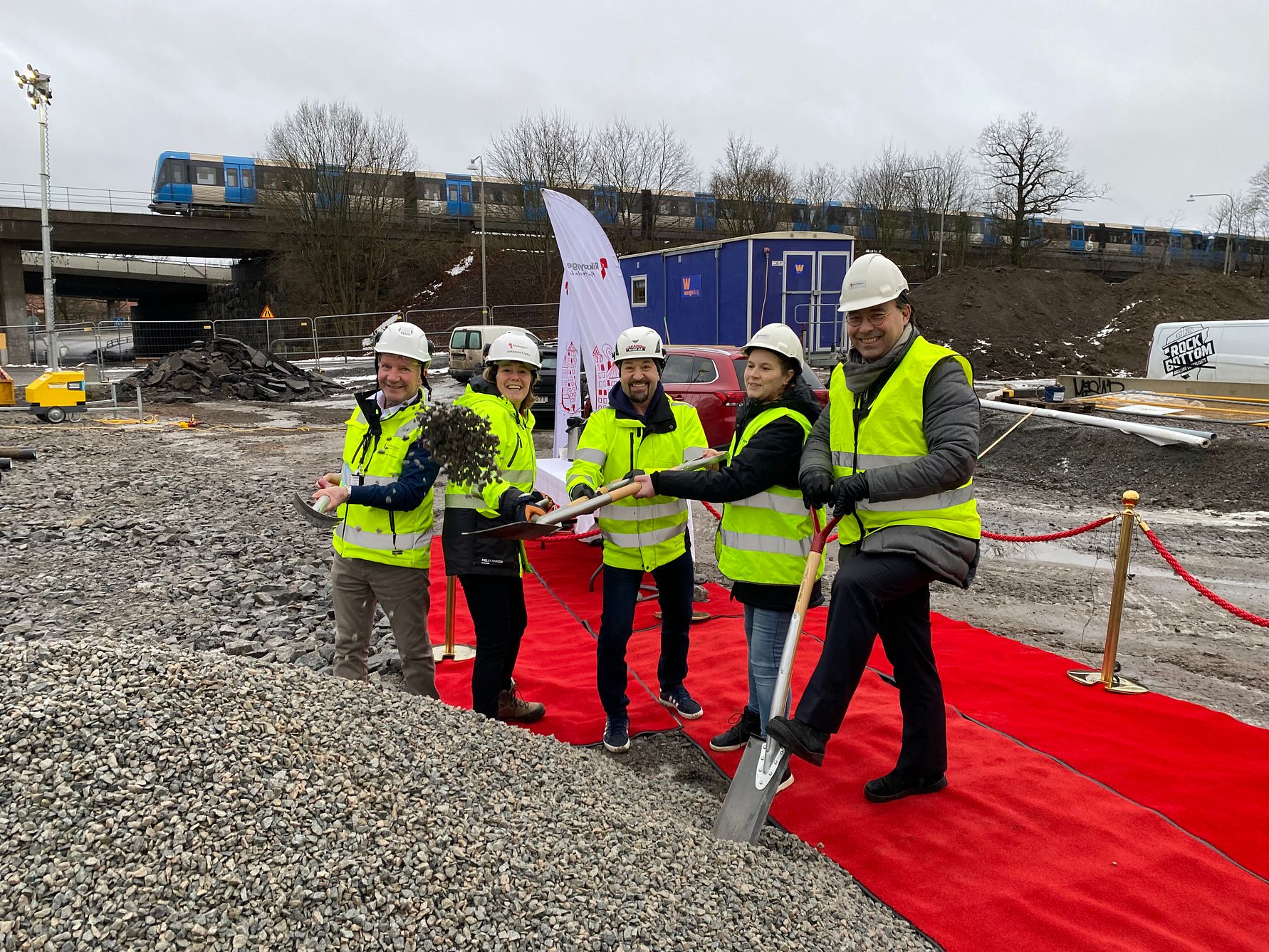 Flera personer i gula säkerhetsvästar och hjälmar deltar i en spadceremoni på en byggarbetsplats med grus och en röd matta. En blå byggbarack och ett passerande tåg syns i bakgrunden