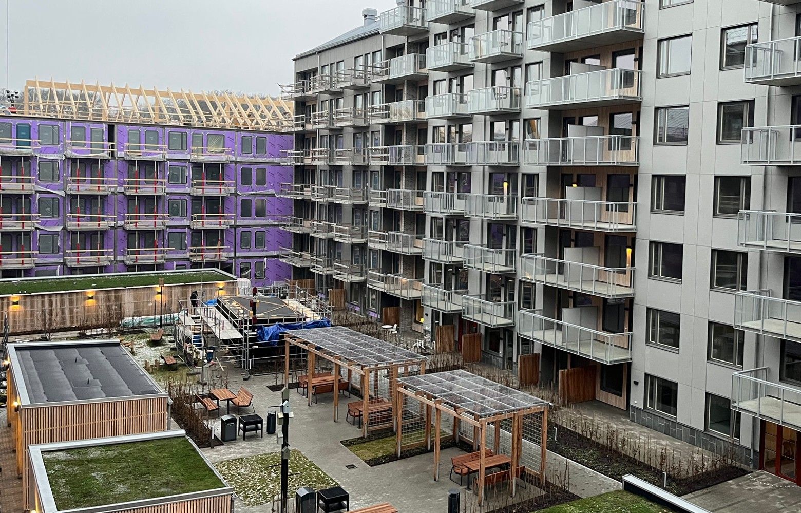 Modern bostadsområde med flerfamiljshus, innergård med pergolor, bänkar och planteringar, samt ett byggprojekt i bakgrunden