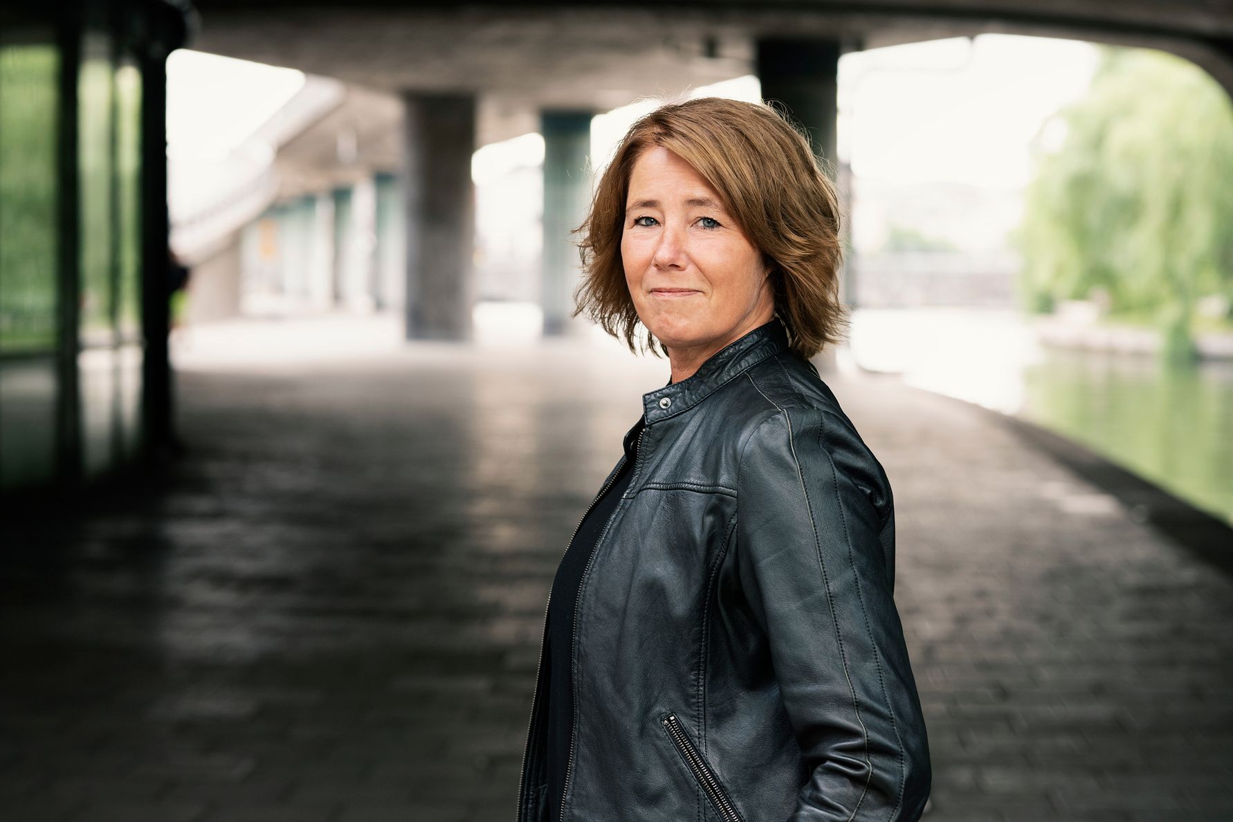 En person med axellångt hår och en svart läderjacka står under en bro med en gångväg och vatten i bakgrunden