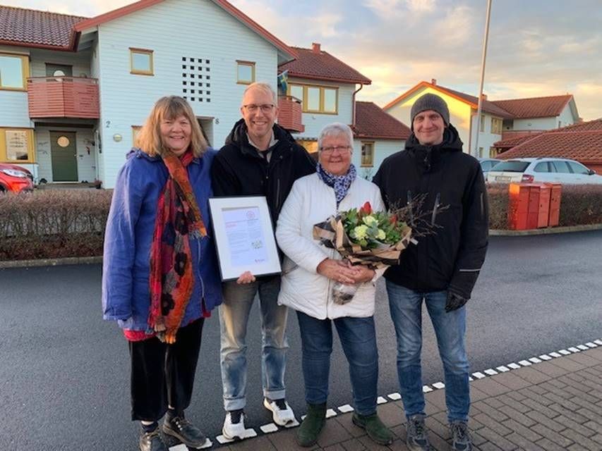 Fyra personer står utomhus på en gata framför hus, varav en håller i en plakett och en annan i en blombukett
