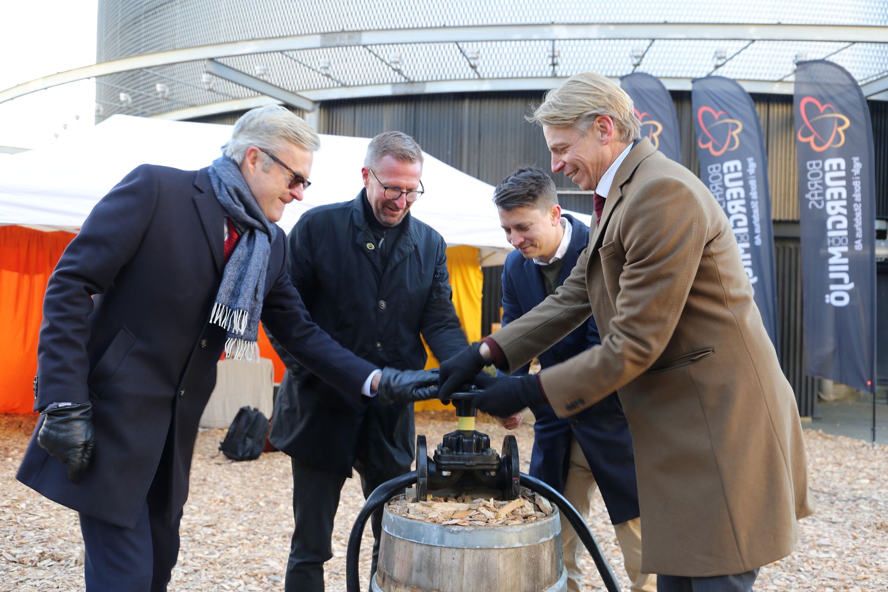 Fyra personer står utomhus runt en träfat och använder en vevmekanism, omgivna av tält och flaggor med logotyper i bakgrunden