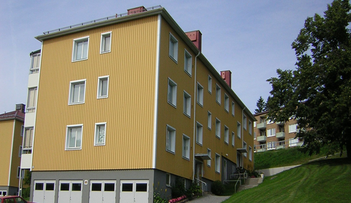 Gul flerfamiljsfastighet med tre våningar, garageportar på bottenplan och omgivning med gräsmatta och träd