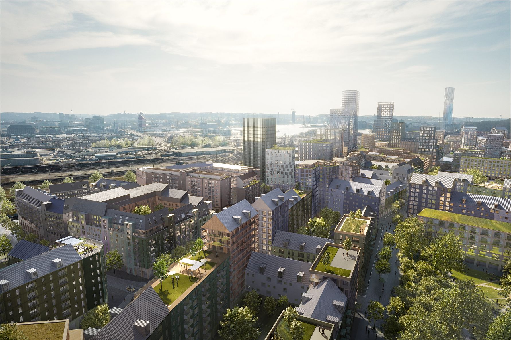 Flygbild över en stadsdel med moderna byggnader, grönområden och en bakgrund med fler höghus och industriområden under en klar himmel