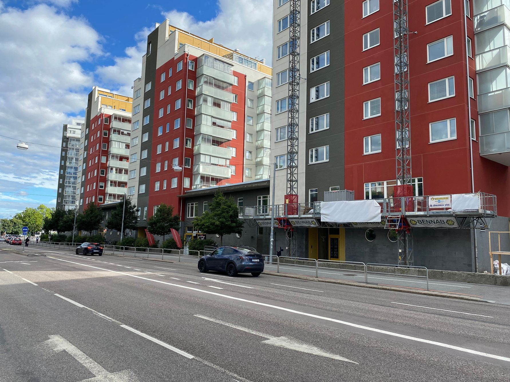 Flerbostadshus med röda och grå fasader längs en gata med parkerade och passerande bilar under en klarblå himmel