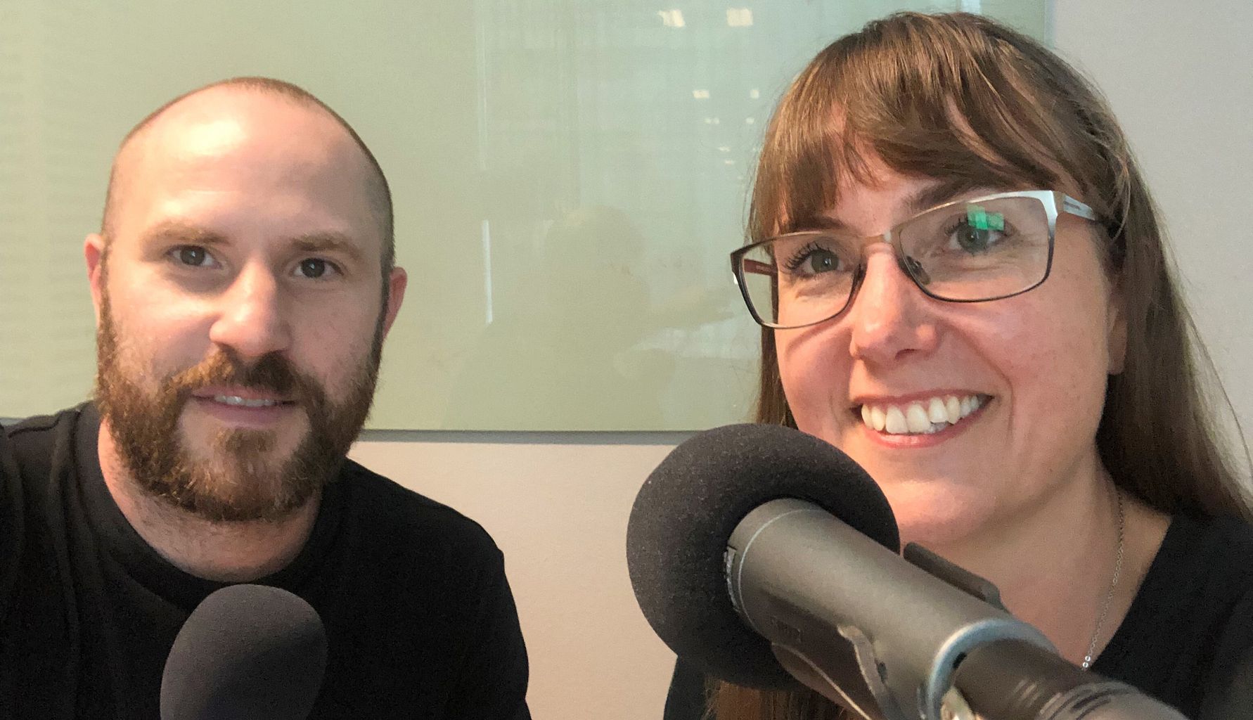 Två personer sitter framför varsin mikrofon i en inspelningsstudio, med bakgrund av en glasvägg och otydliga konturer av ett kontorsutrymme