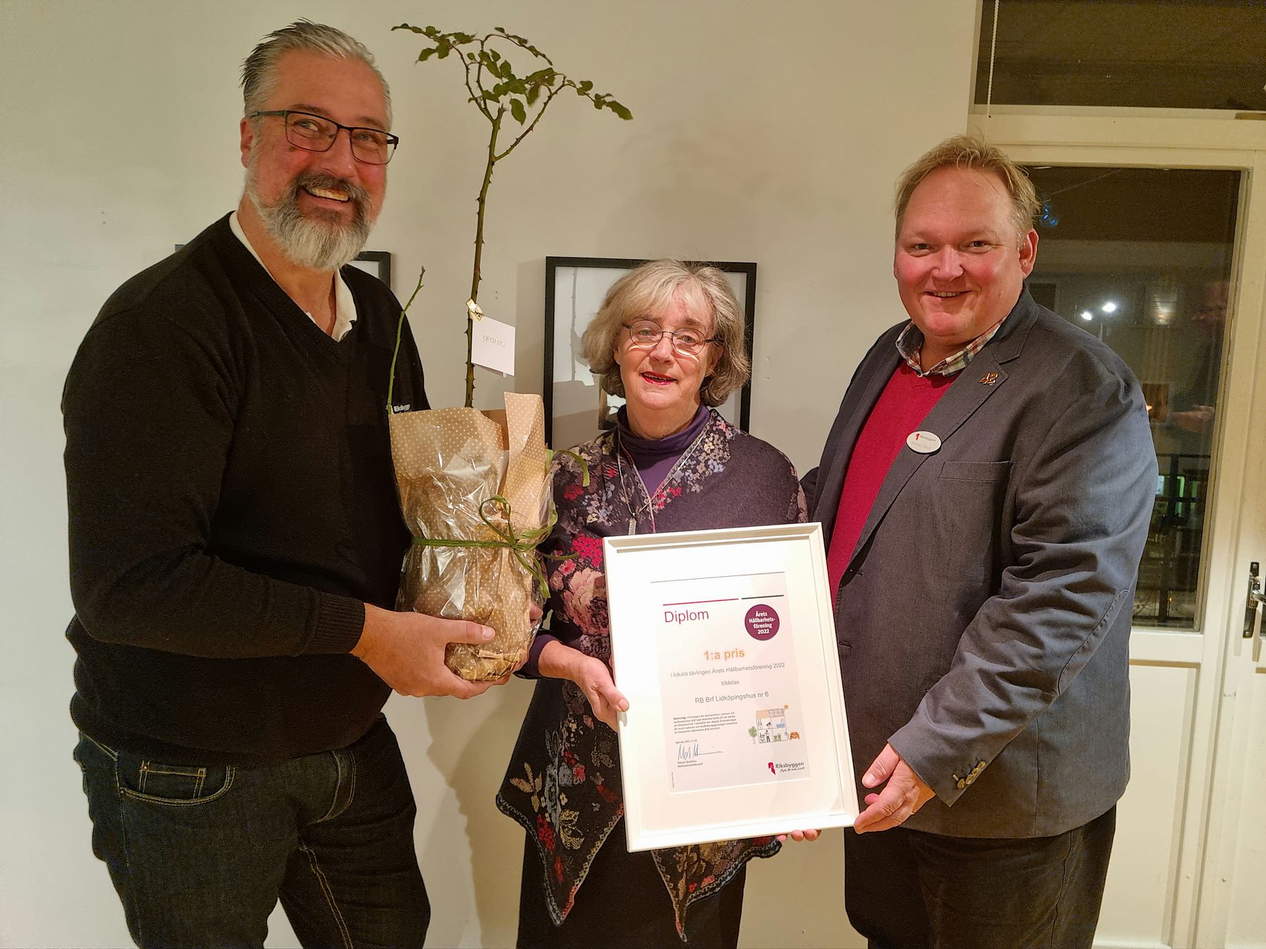 Tre personer står inomhus, och en av dem håller ett diplom i en ram medan en annan håller en inslagen planta