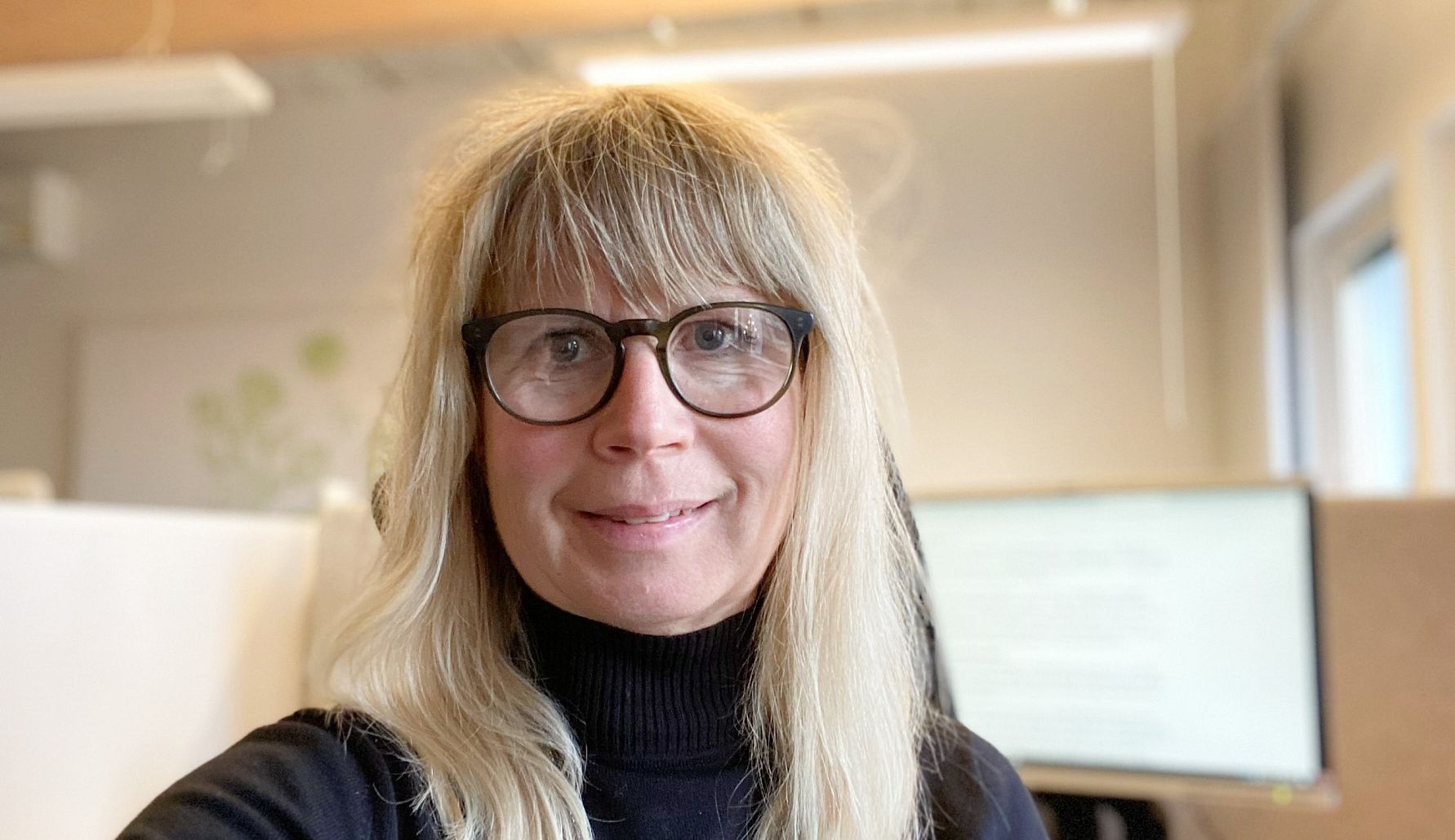 En person med långt blont hår och svart polotröja sitter i ett kontorsutrymme med suddiga skärmar och ljusa trädetaljer i bakgrunden