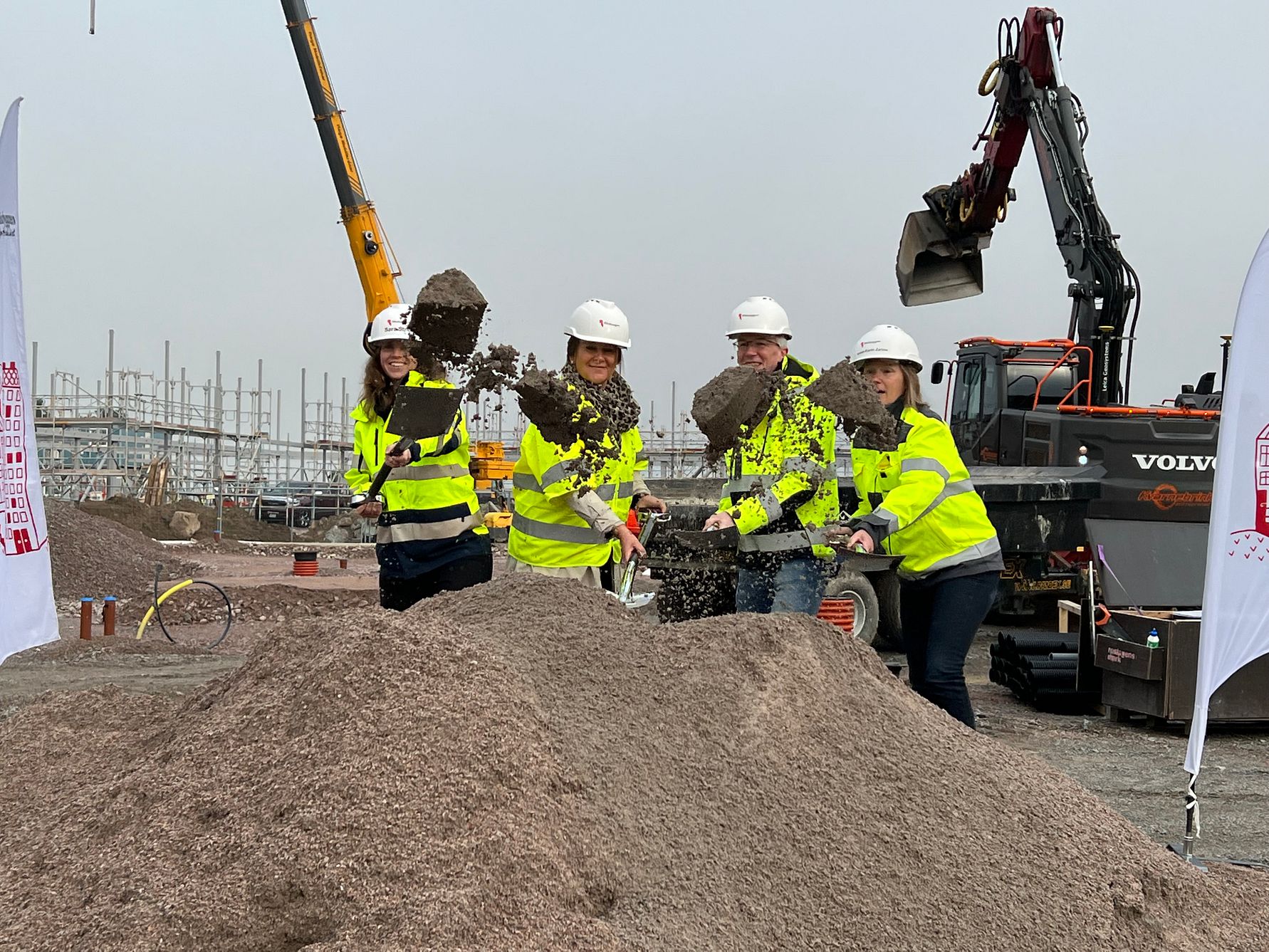 Fyra personer i gula reflexjackor och vita hjälmar skyfflar grus vid en byggarbetsplats med grävmaskiner och lyftkran i bakgrunden