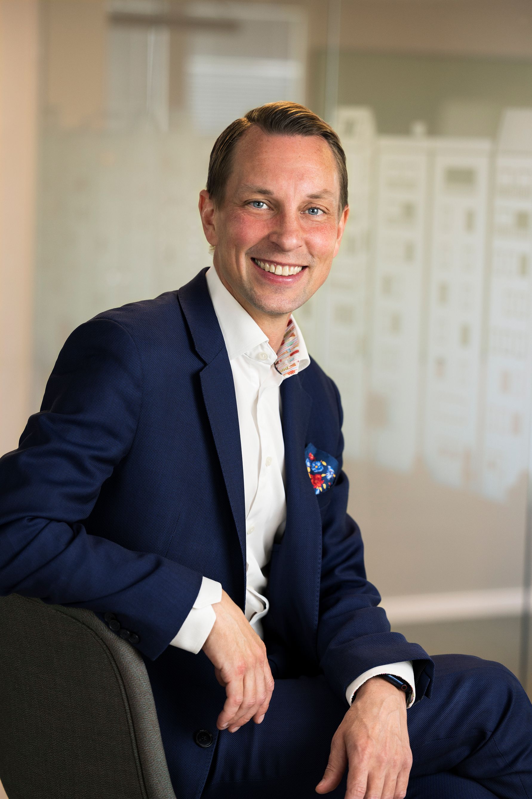 En person i kostym sitter på en stol med ena handen på armstödet, bakgrunden visar en suddig kontorsmiljö med glaspaneler och ett mönster av byggnader