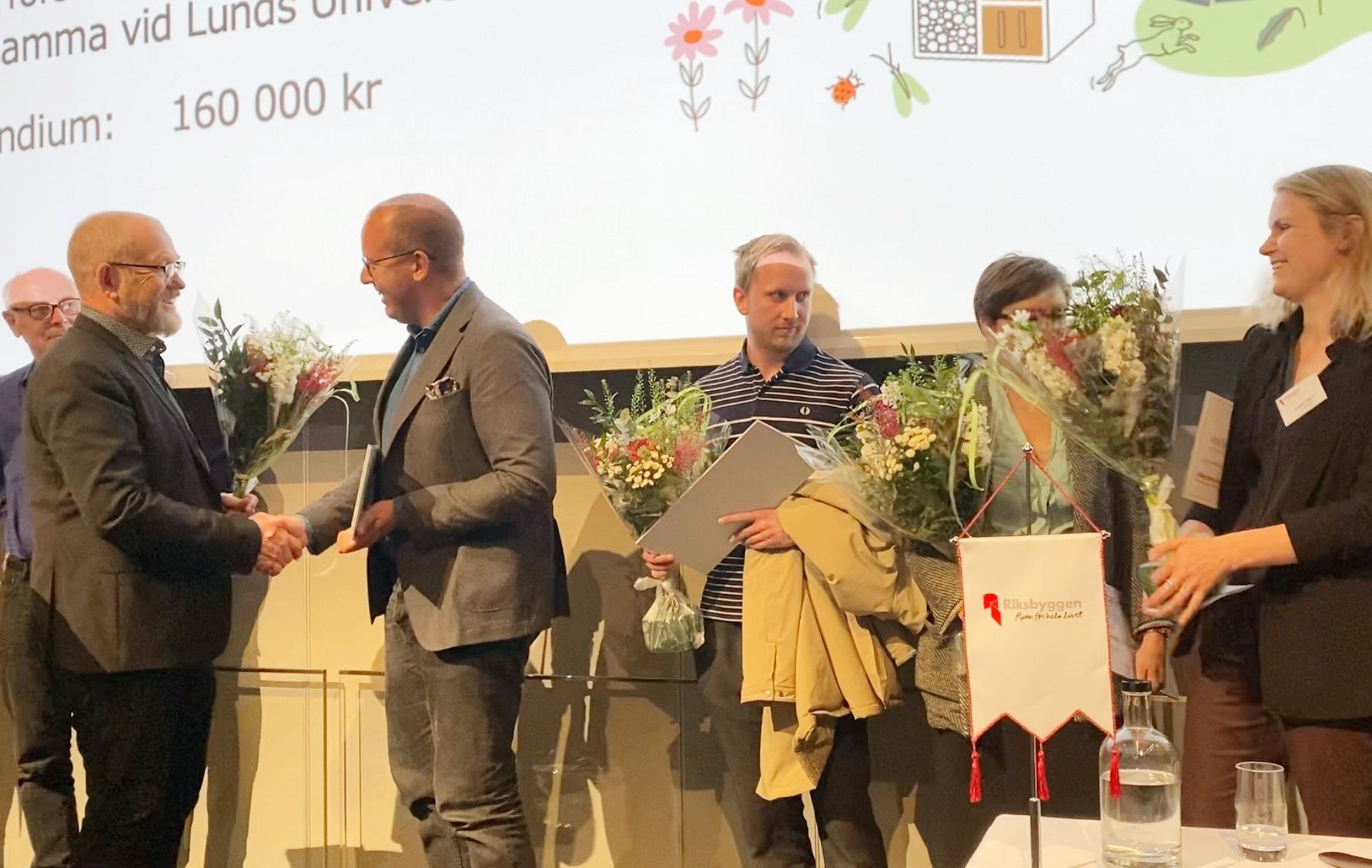En grupp människor på en scen som får utmärkelser med blommor och dokument, framför en projekterad presentation