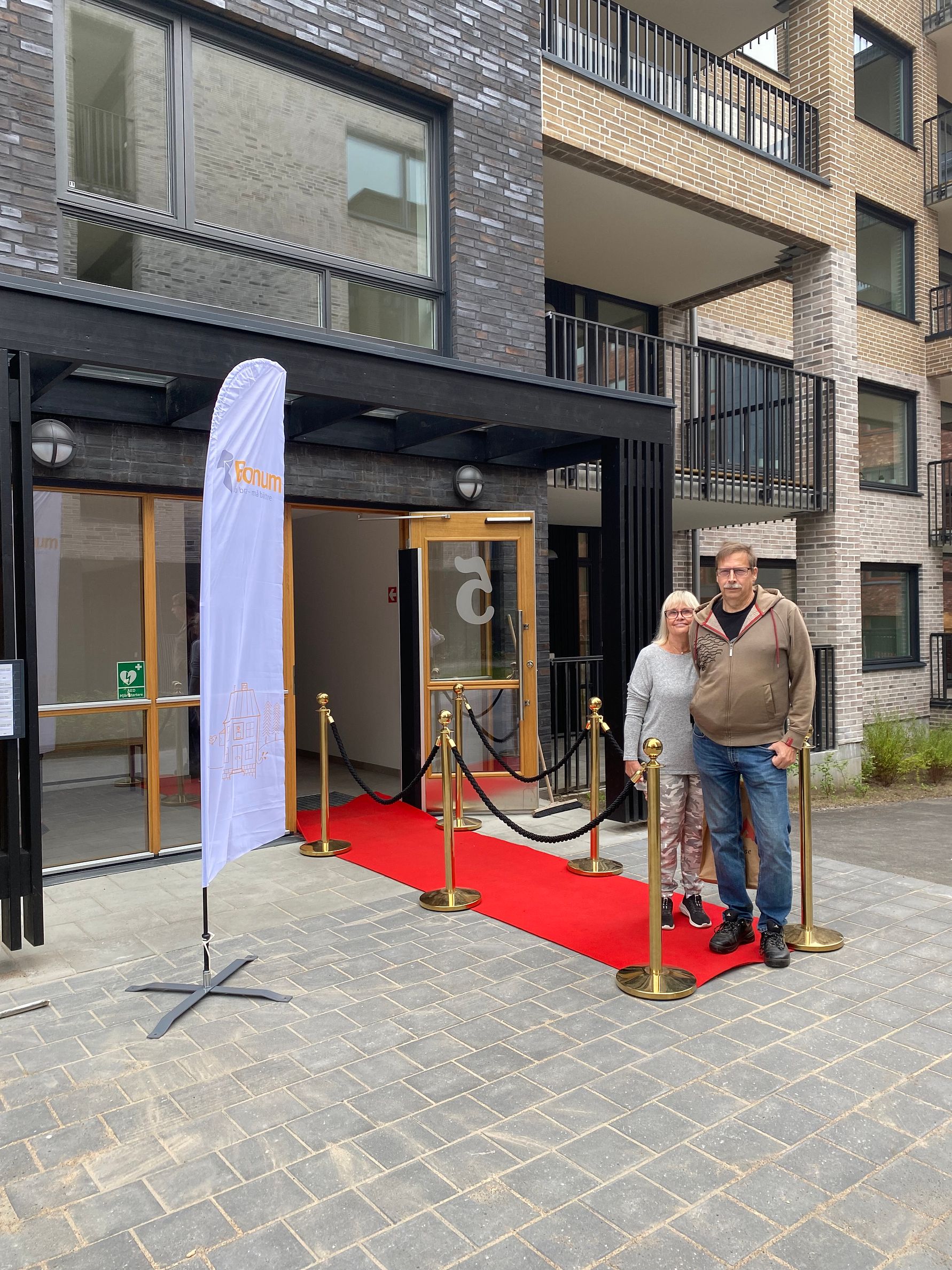 Två personer står på en röd matta framför ingången till en modern byggnad med svarta och ljusa fasaddetaljer