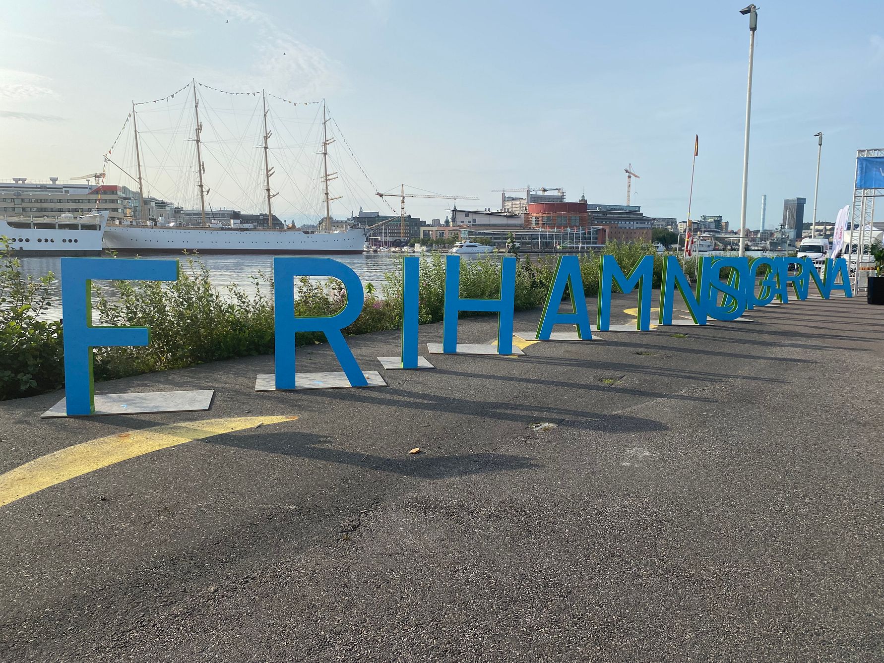 Skylt med texten "Frihamnsbornas" i blå bokstäver vid en kaj med fartyg och byggnader i bakgrunden