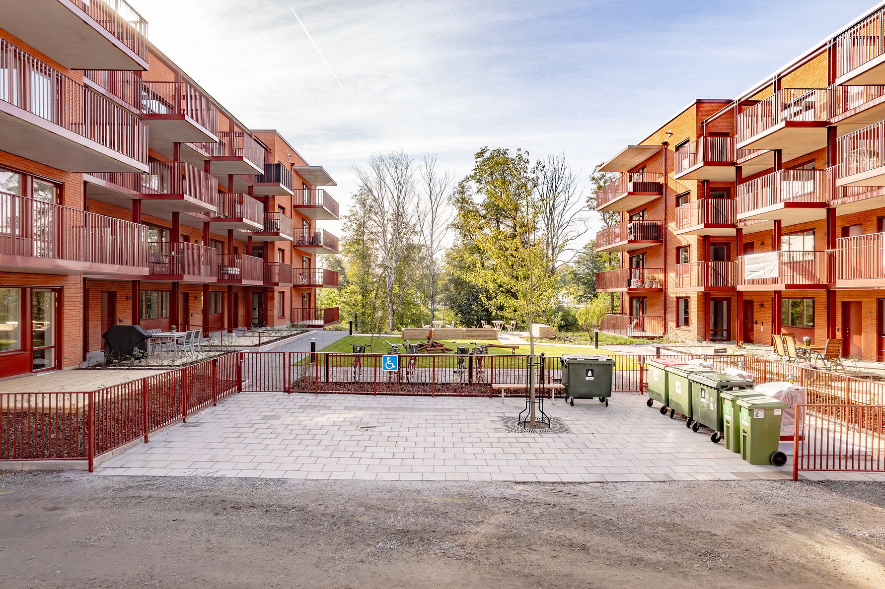 Lägenhetsbyggnader med röda balkonger på vardera sida, en innergård med gräsmatta och träd samt sopkärl på en stenlagd yta