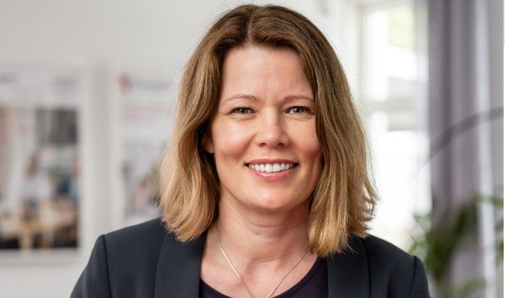 Kvinna med axellångt blont hår i en mörk kavaj, stående inomhus med suddig bakgrund bestående av fönster och affischer