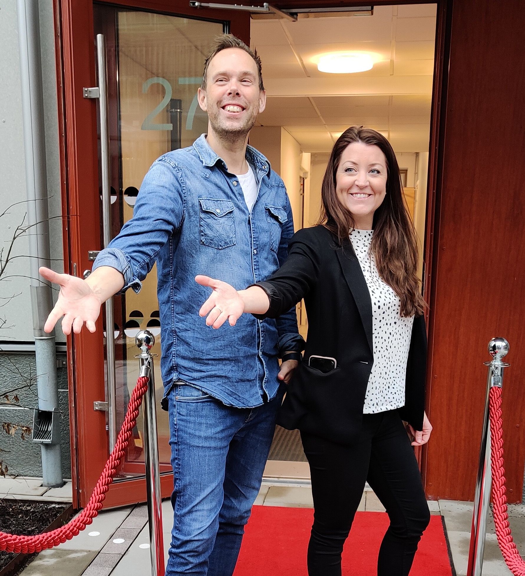 Två personer står framför en dörr med röda repstolpar och en röd matta. Den ena bär jeans och jeansskjorta, och den andra en svart kavaj med vit prickig topp