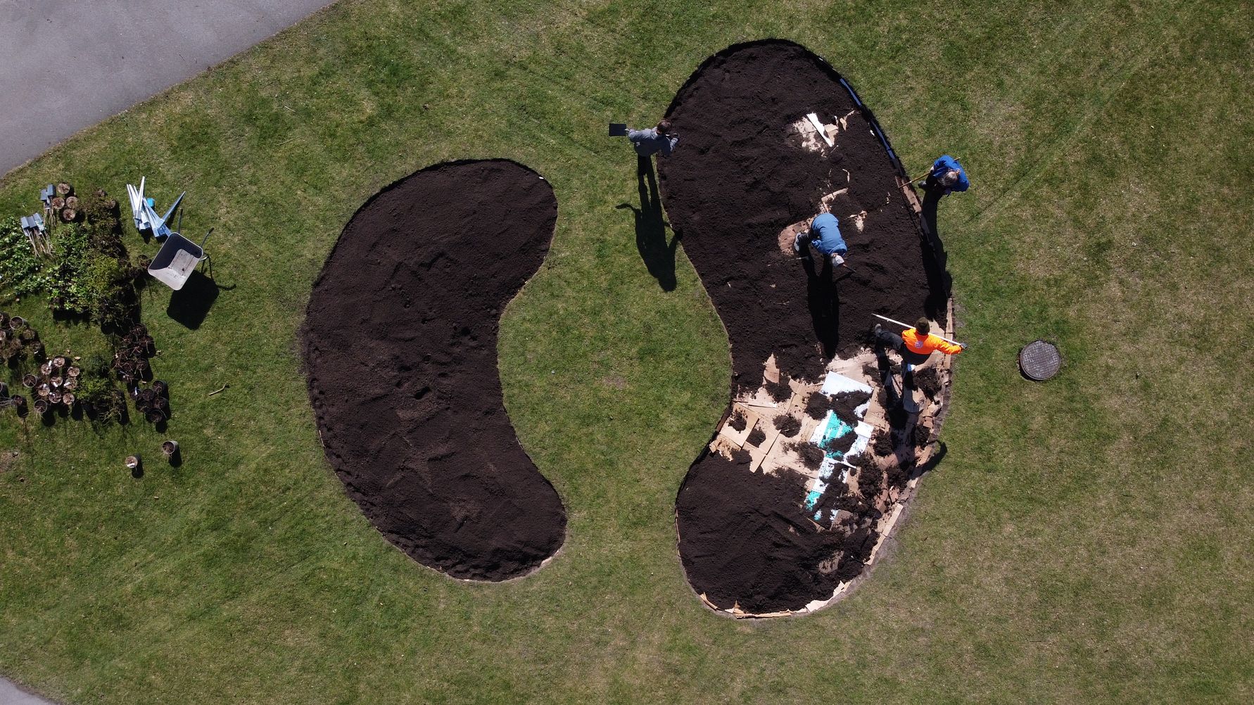 Luftbild av två jordytor formade som böjda ovaler på en gräsmatta, med personer som arbetar med marken