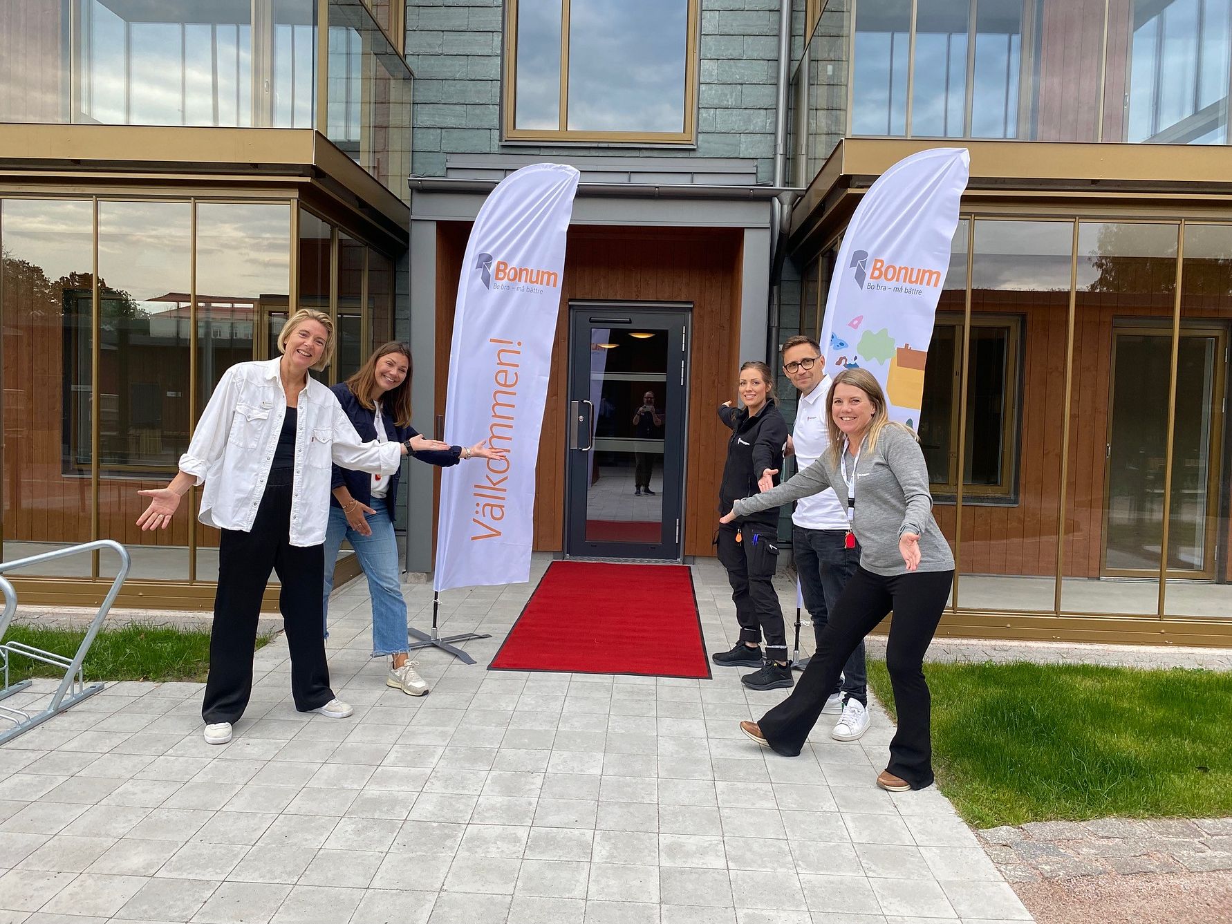En grupp människor står framför en entré till en byggnad med glasfasad. Två flaggor med texten "Bonum" och en röd matta syns vid ingången