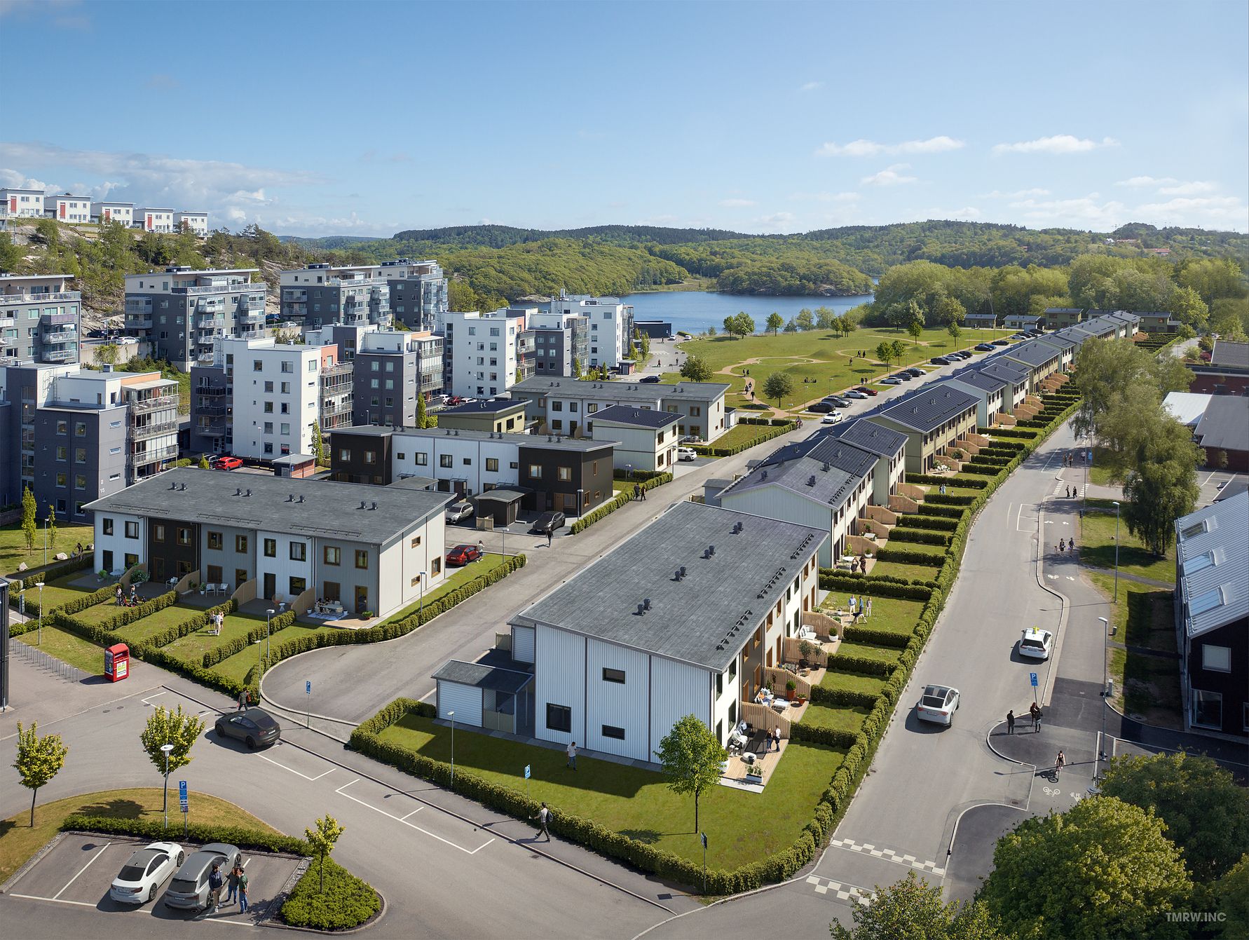 Bostadsområde med moderna flerfamiljshus och radhus, omgivna av grönska och en sjö i bakgrunden. Klarblå himmel och soligt väder
