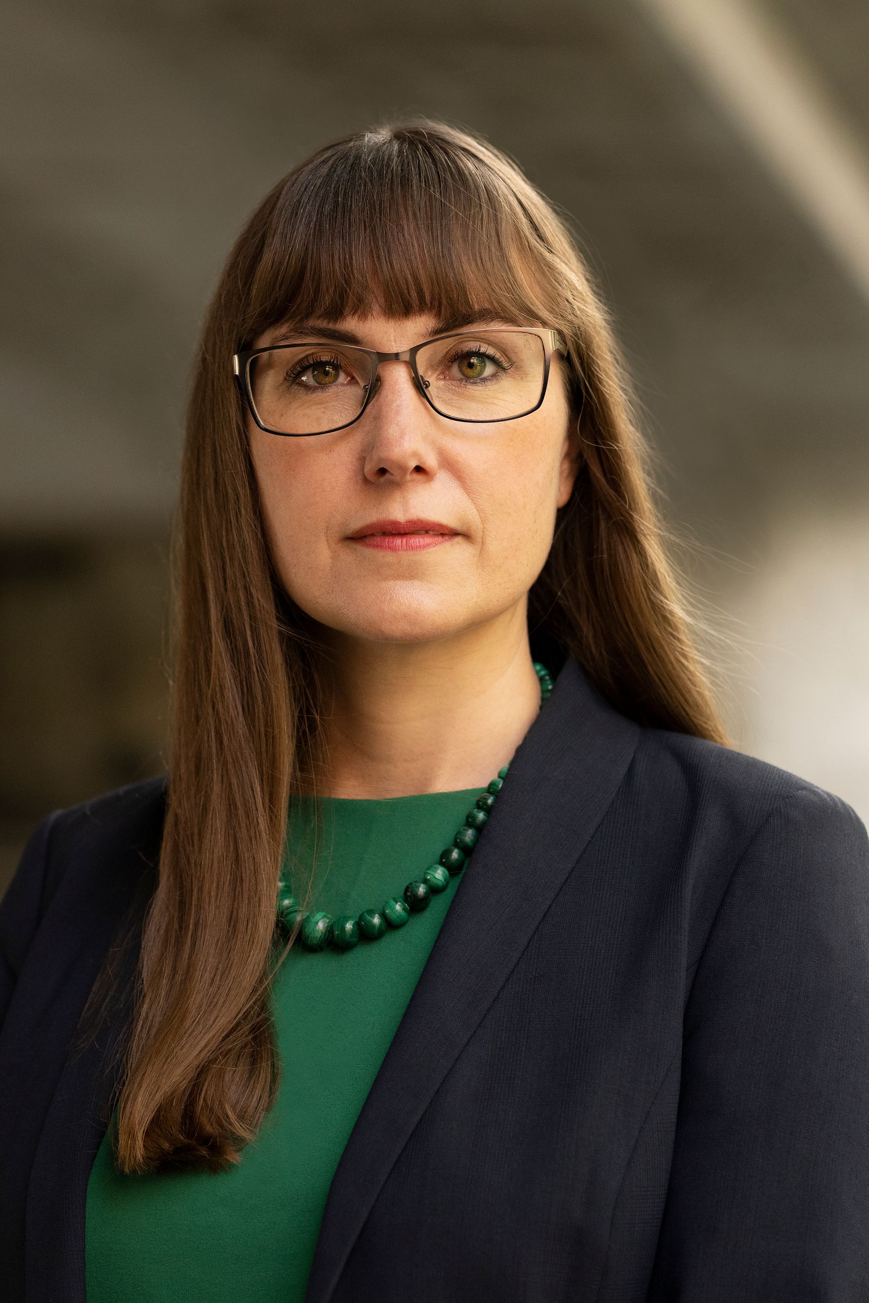 En kvinna med långt brunt hår, klädd i en grön topp och en mörkblå kavaj, bär ett grönt pärlhalsband