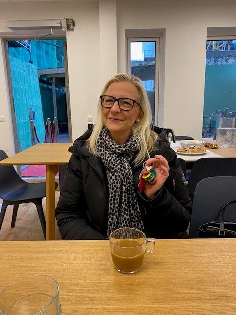 En person sitter vid ett träbord med en kopp dryck framför sig, håller i en liten samling färgglada föremål, och är klädd i en svart jacka och leopardmönstrad halsduk. I bakgrunden syns fönster och en utomhusmiljö