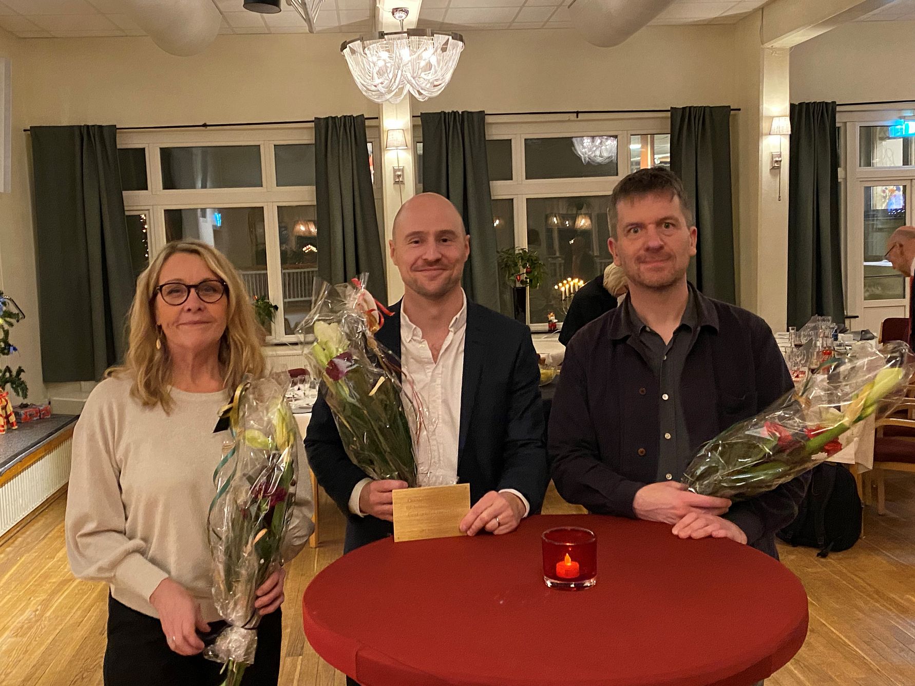 Tre personer står vid ett runt bord med röda dukar, hållandes blombuketter och ett kuvert. Bakgrunden visar ett rum med fönster och gardiner, belysning och dekorationer