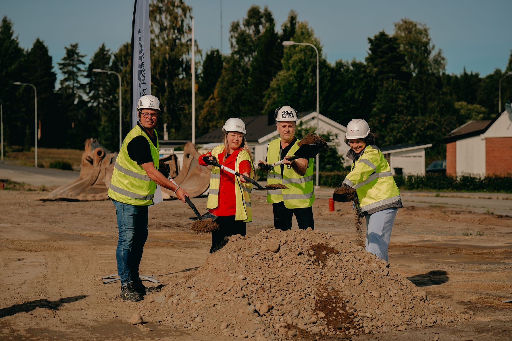 Fyra personer i reflexvästar och hjälmar som använder spadar för att gräva i en jordhög på en byggarbetsplats, med träd och byggnader i bakgrunden