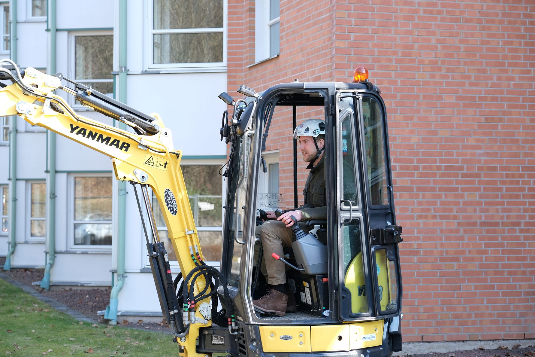 En person kör en gul minigrävare av märket Yanmar framför en byggnad med tegelvägg och fönster