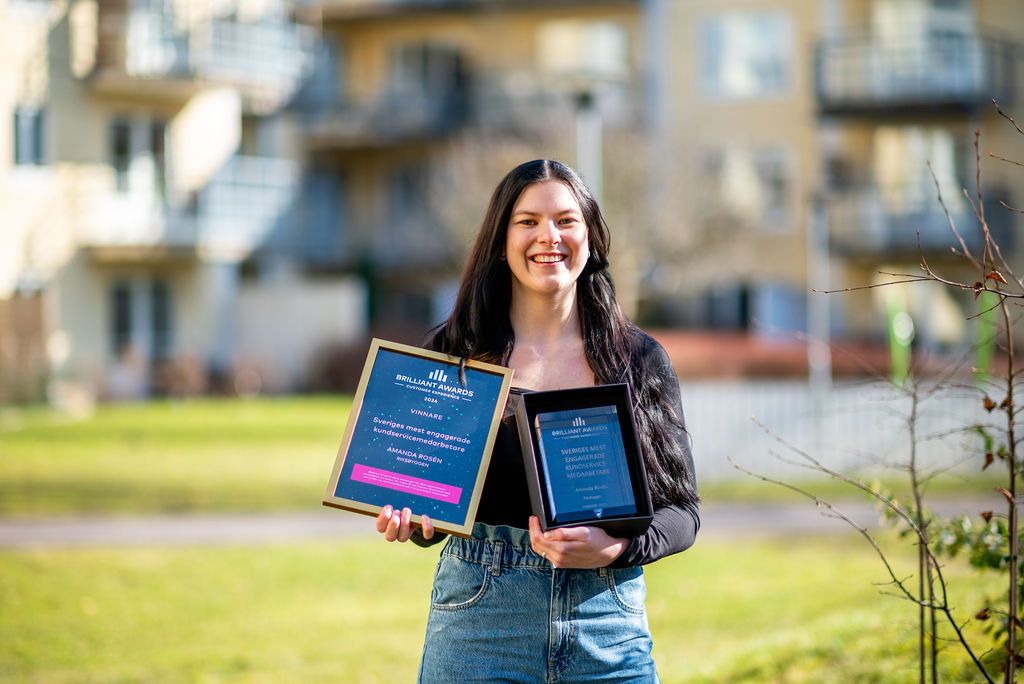 Kvinna med långt mörkt hår håller två diplom, stående utomhus med bostadshus och grönska i bakgrunden