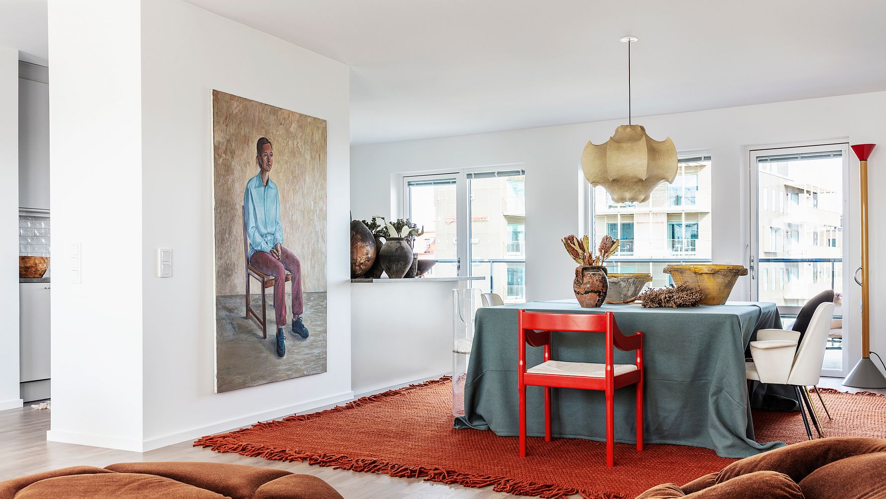 Ett modernt vardagsrum och matplats med vitmålade väggar, en stor matta i terrakottafärg, ett bord med en grå duk, röda stolar och dekorativa vaser