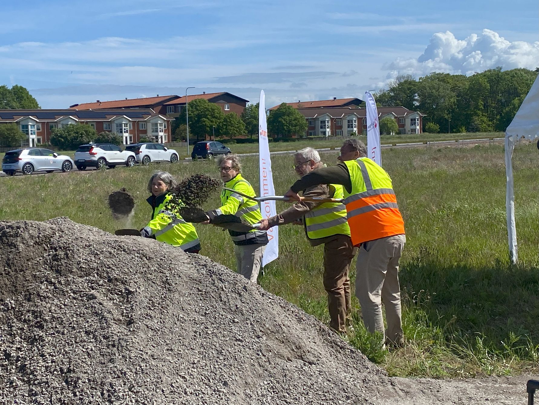Flera personer i reflexvästar skyfflar grus vid ett grönområde, med byggnader och bilar i bakgrunden