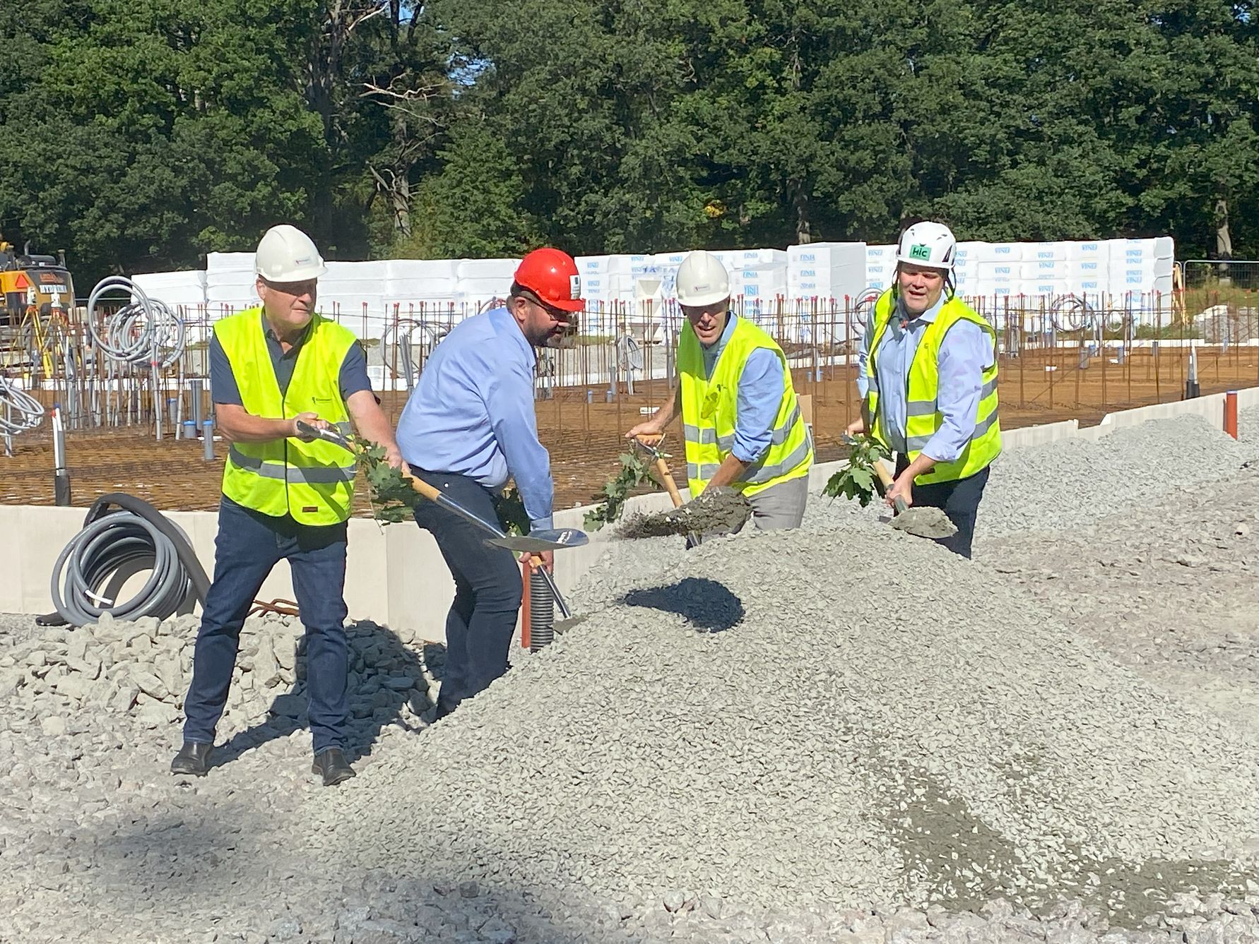 Byggarbetsplats med fyra personer i skyddsvästar och hjälmar som arbetar med en grushög. Bakgrunden visar byggmaterial och träd