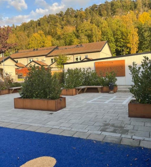 Utomhusmiljö med stenlagd gård, planteringslådor och bänkar framför byggnader, omgiven av träd och en skog i bakgrunden