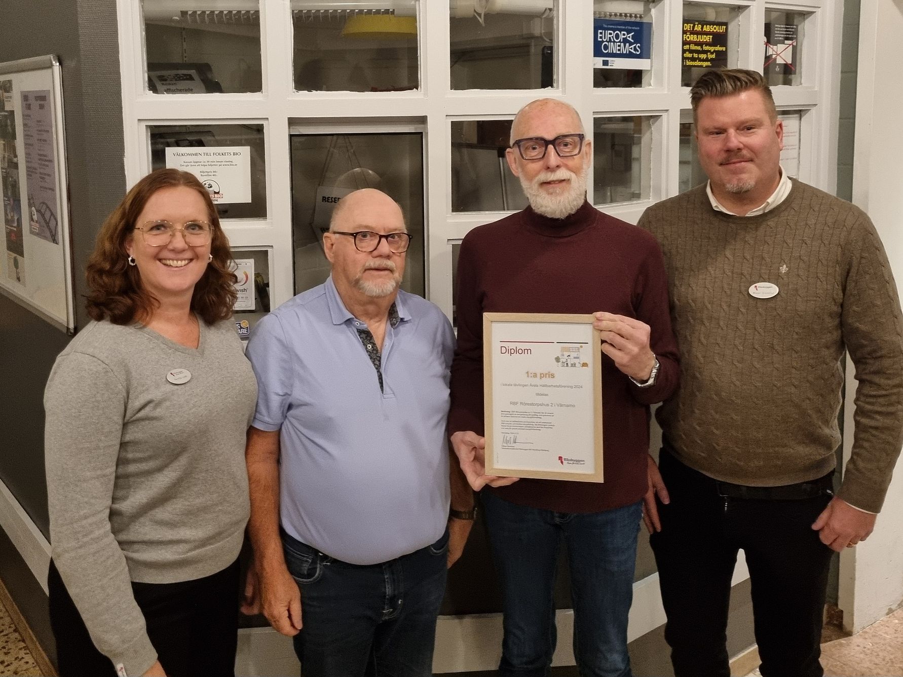 Fyra personer står framför en vägg med fönster, varav en person håller ett diplom. Bakgrunden visar anslag och skyltar på väggarna