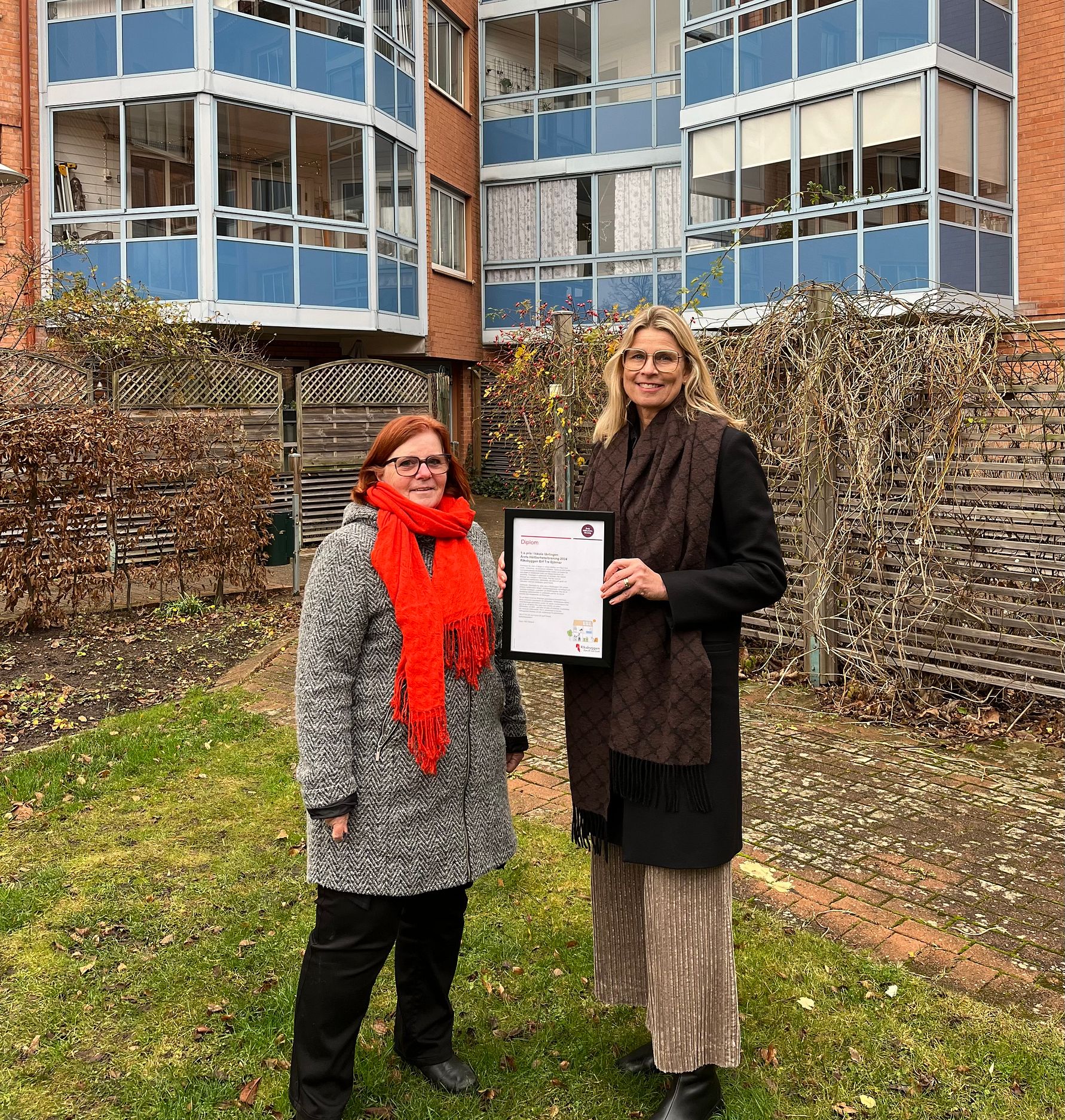 Två personer står på en gräsmatta framför en byggnad med stora fönster och balkonger, en av dem håller ett inramat dokument